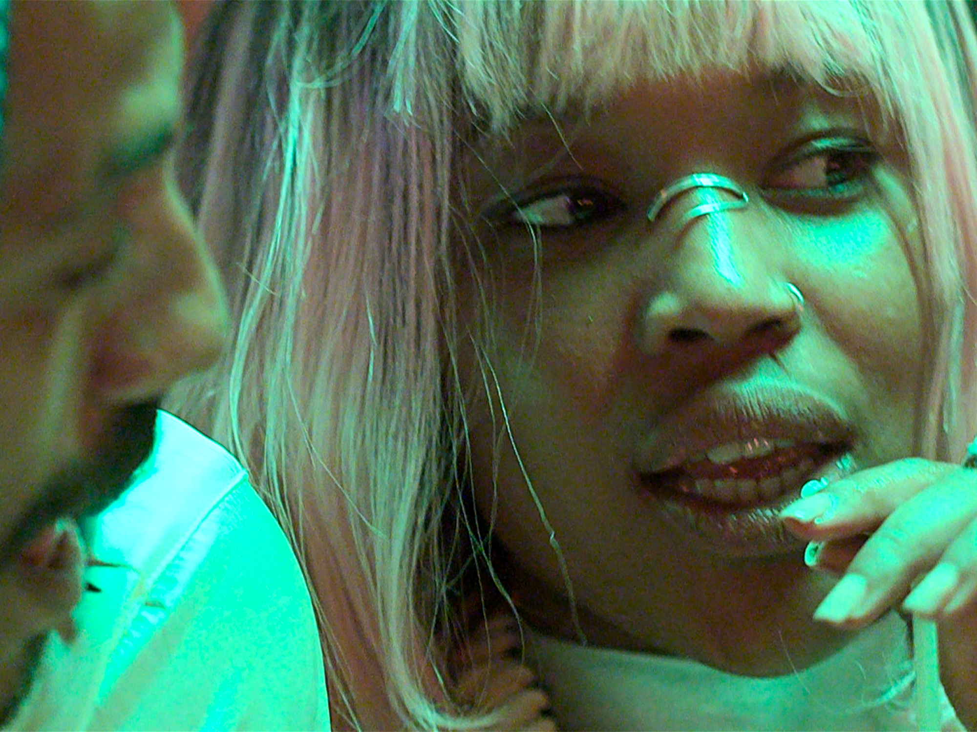 Woman with blonde hair and fringe in bright green and blue lighting, hand near face, close-up portrait with dramatic colour cast.