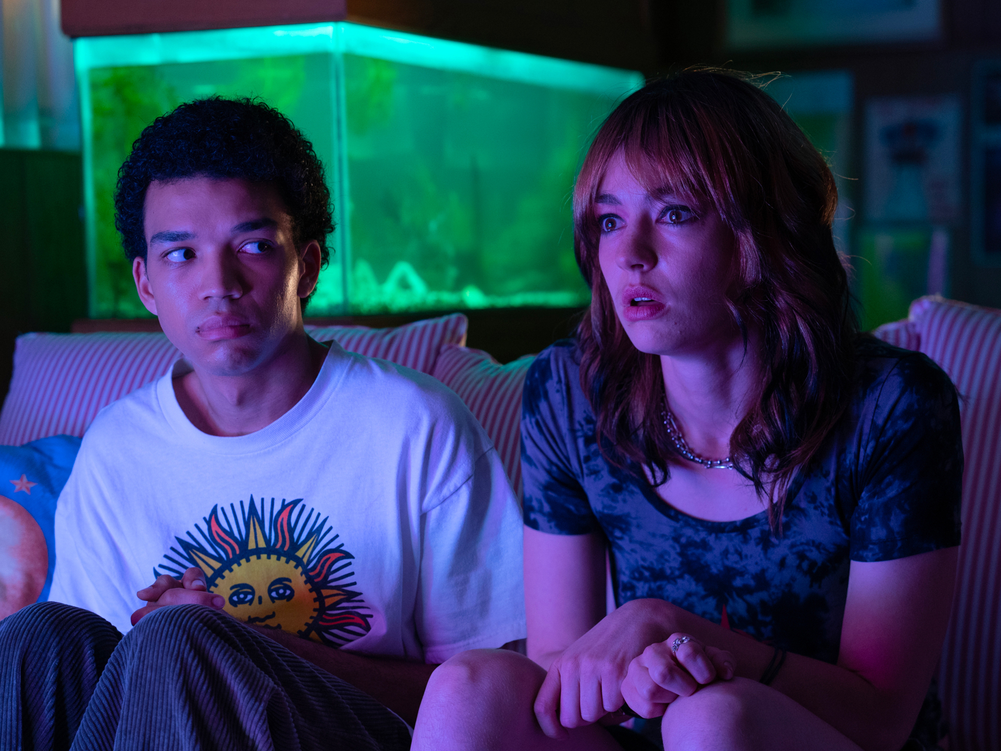 A young man and woman sitting on a couch, surrounded by a neon green lighting.