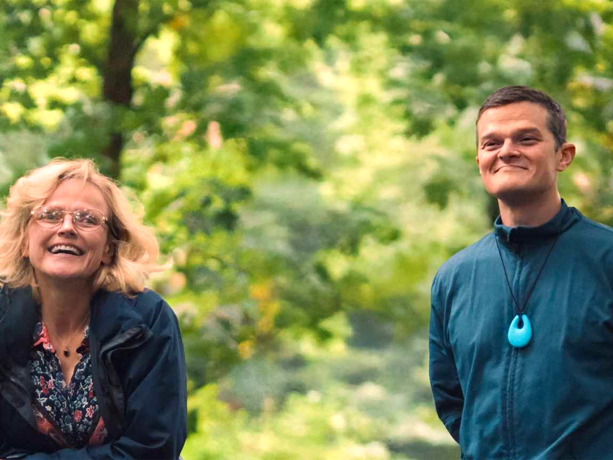 Woman with blonde hair and glasses in dark jacket laughing beside man in teal jumper, both standing outdoors with green foliage.