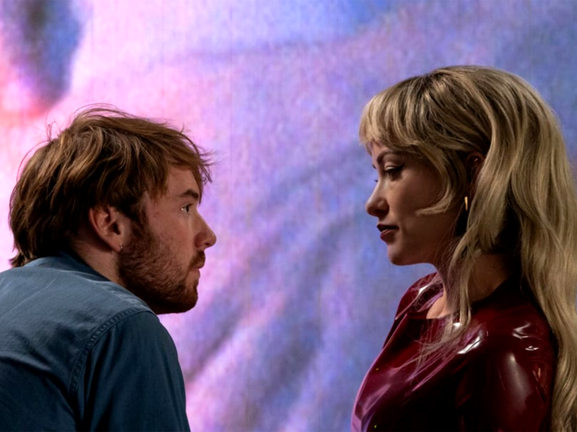 Man with brown hair and beard in blue shirt facing blonde woman in red leather jacket against purple and pink cloudy background.