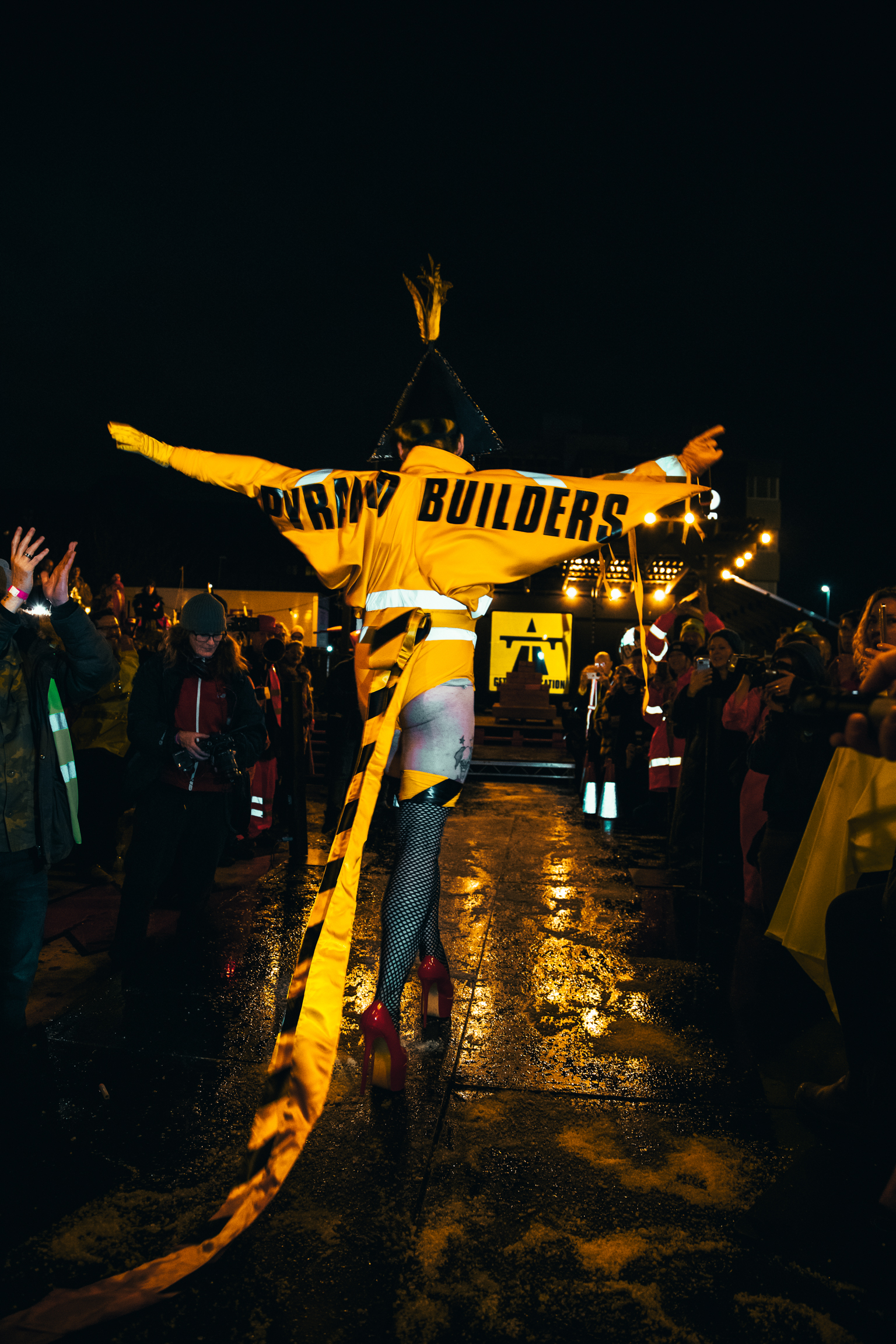 Person holding large yellow banner reading "BUILDERS" with arms outstretched amongst crowd on wet street at night with lights.