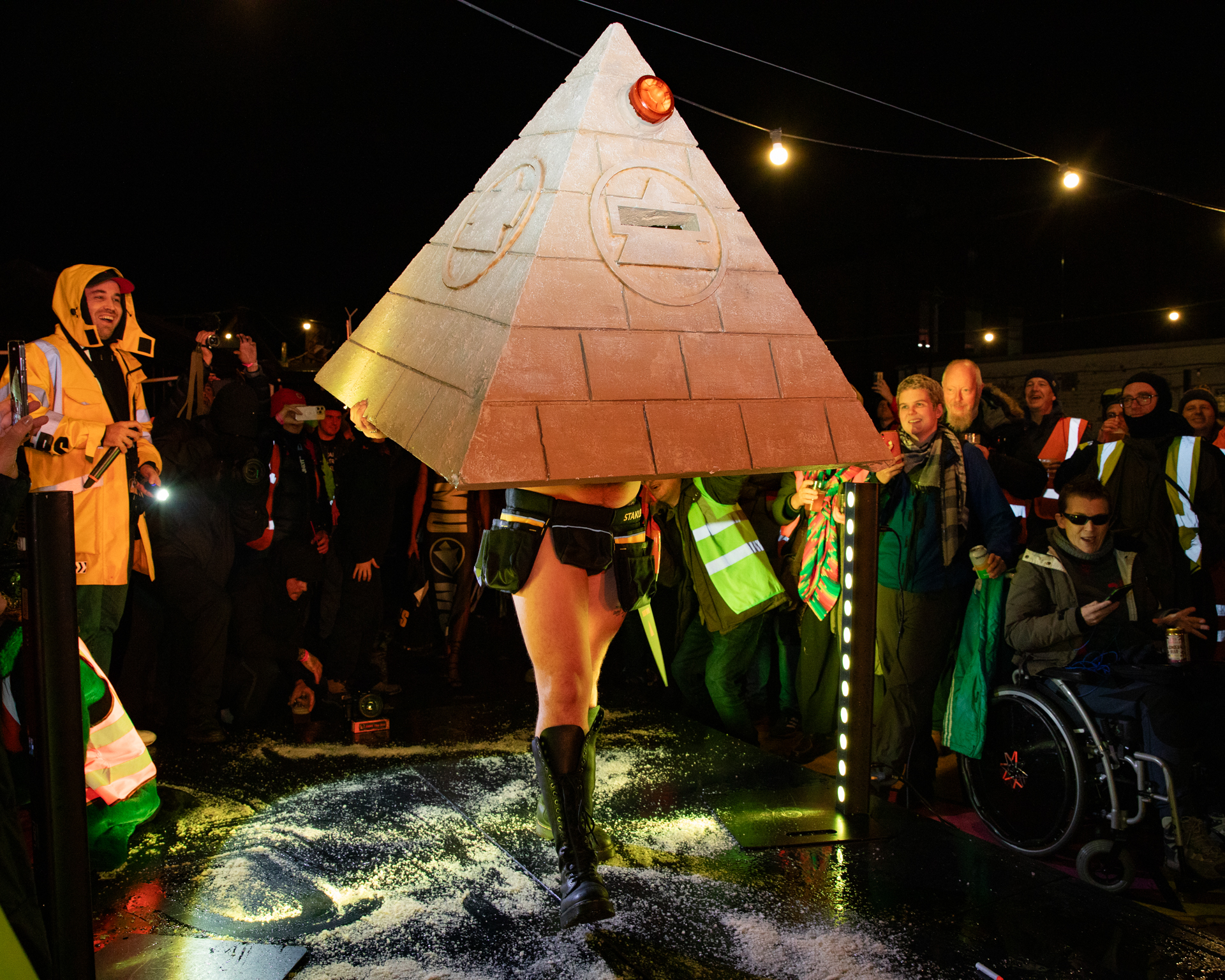 Person wearing large triangular pyramid costume with brick pattern at night-time outdoor event with crowd and string lights overhead.
