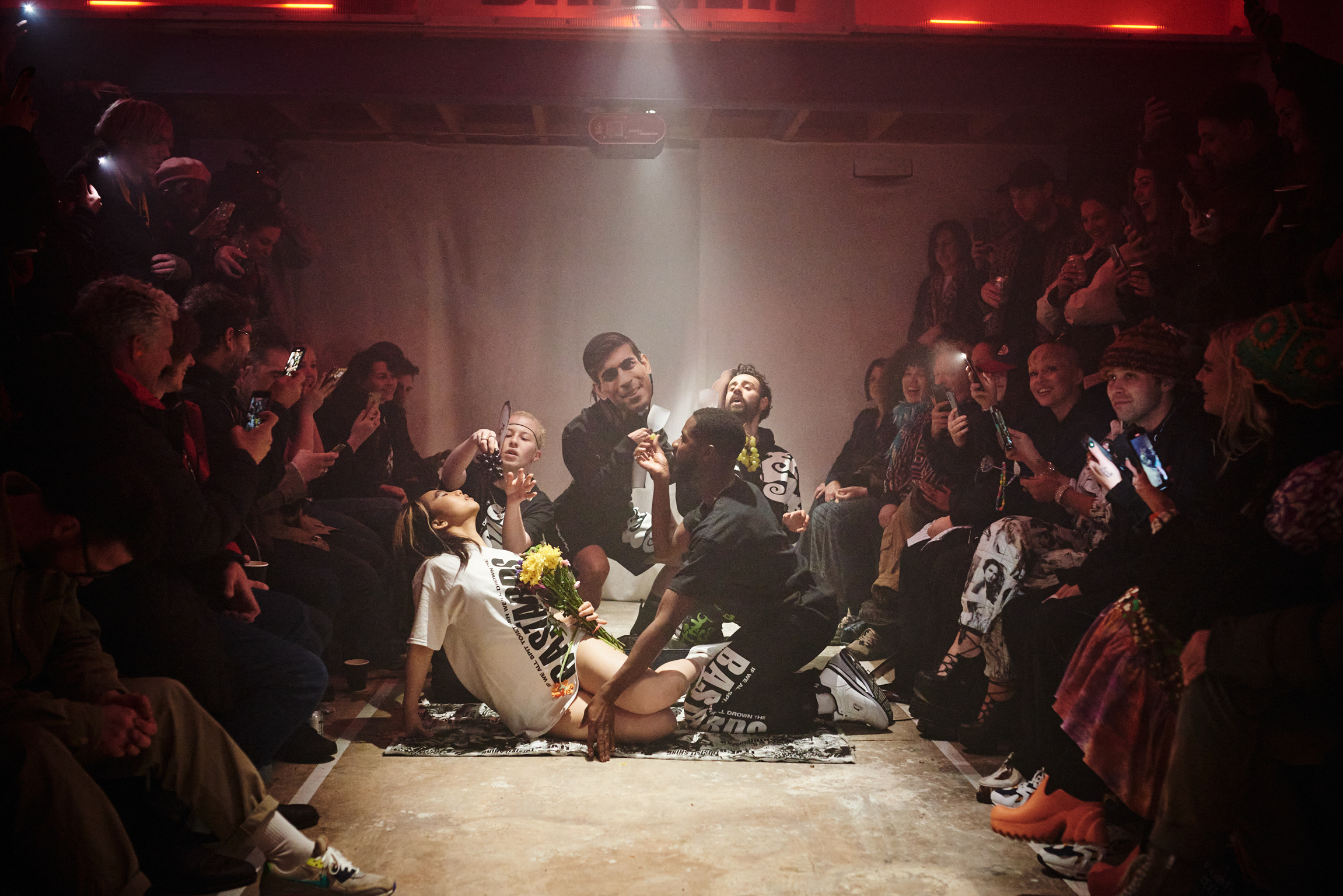 Breakdancers performing in centre of circle formed by seated audience, dramatic overhead lighting, red neon lights above.