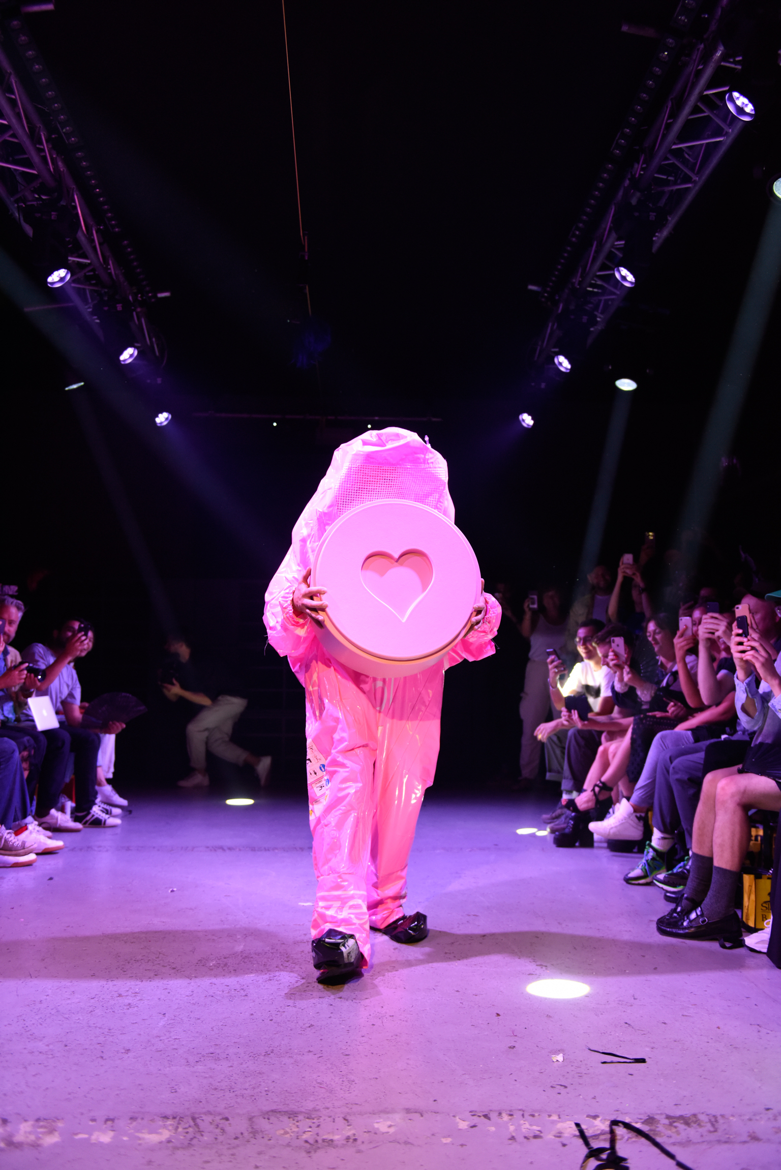 Person in bright pink hooded outfit with heart-shaped face opening on runway, surrounded by seated audience under purple stage lighting.