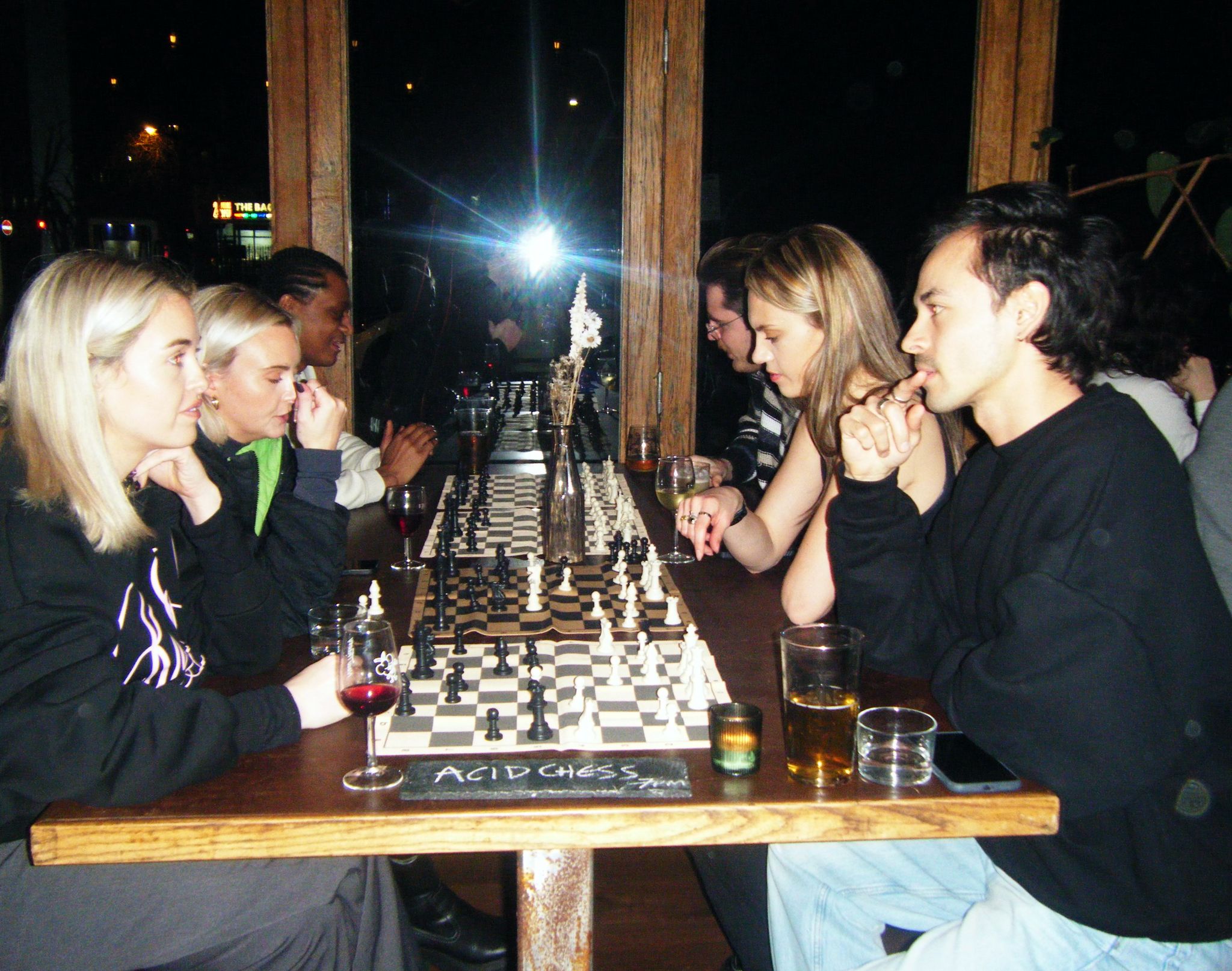 Group of people gathered around wooden table with multiple chess boards, drinks visible, indoor setting with warm lighting.