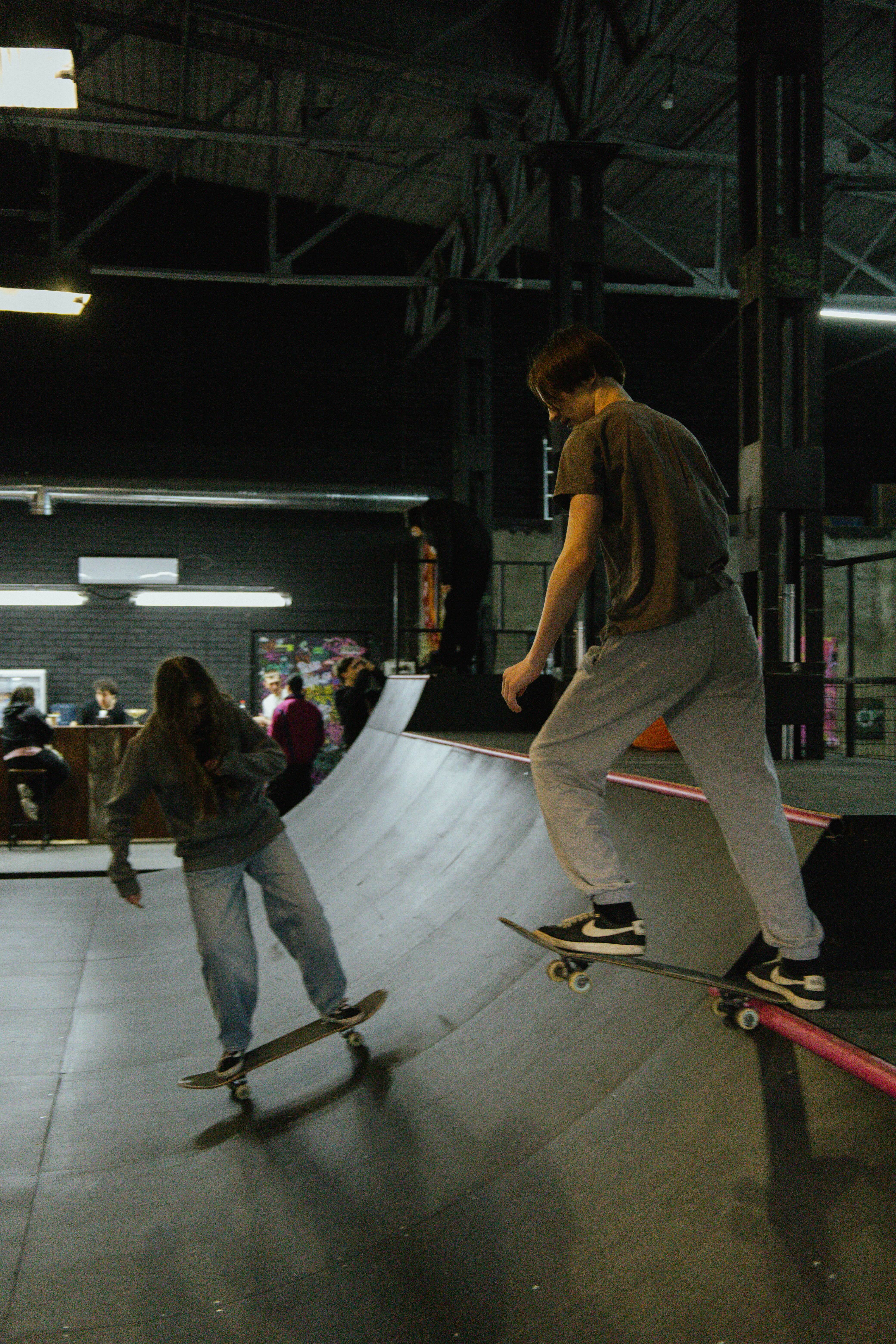 Skateboarders on ramp in indoor skate park, dark industrial setting with people in background.