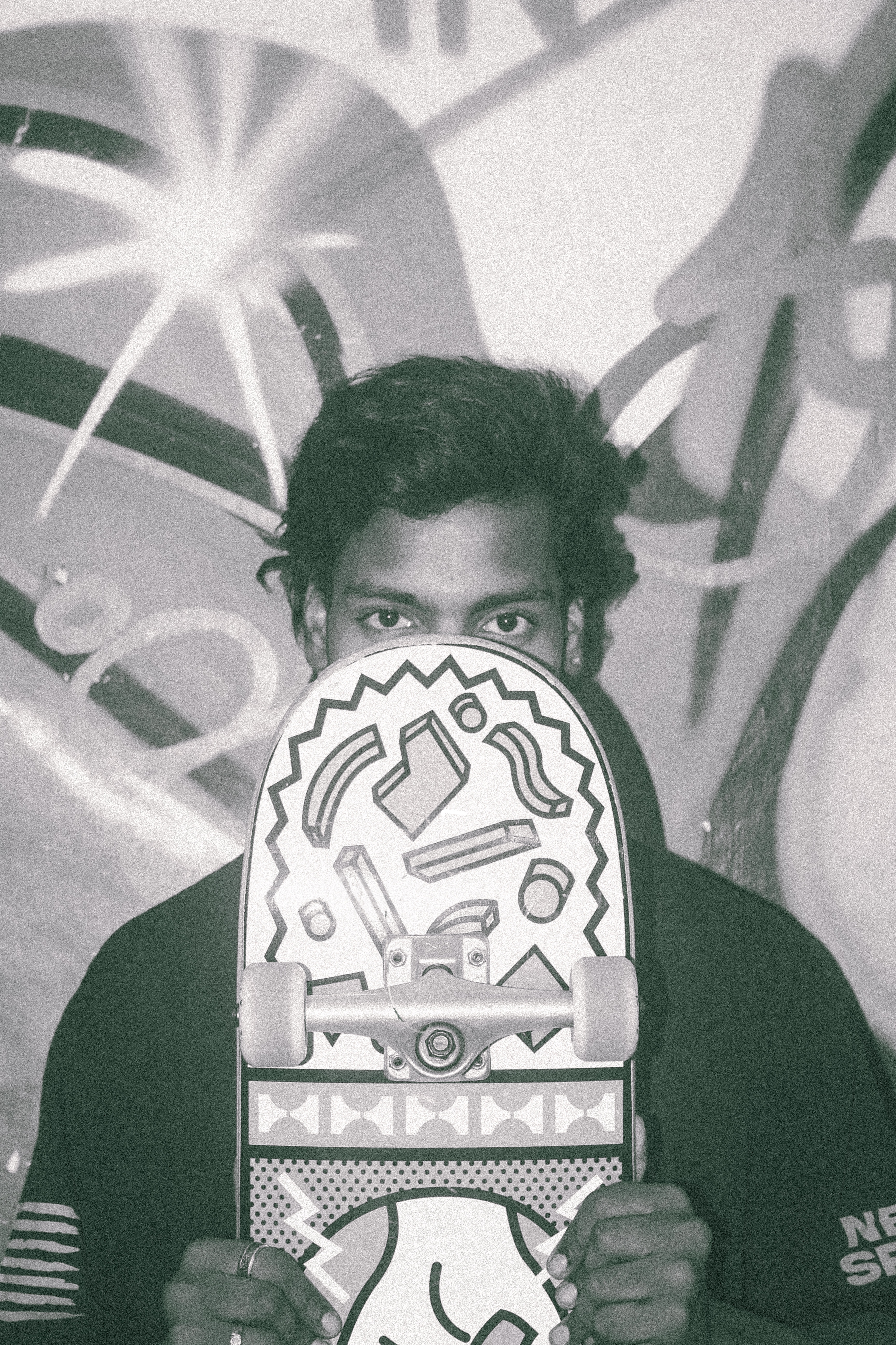 Person holding decorative mask with geometric patterns and tribal-style designs, covering lower face. Graffiti visible on wall behind.