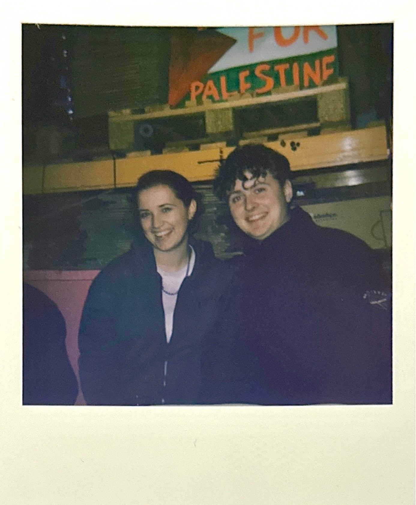 Two people smiling in dark jackets, standing in front of yellow school bus with "PALESTINE" sign visible above.