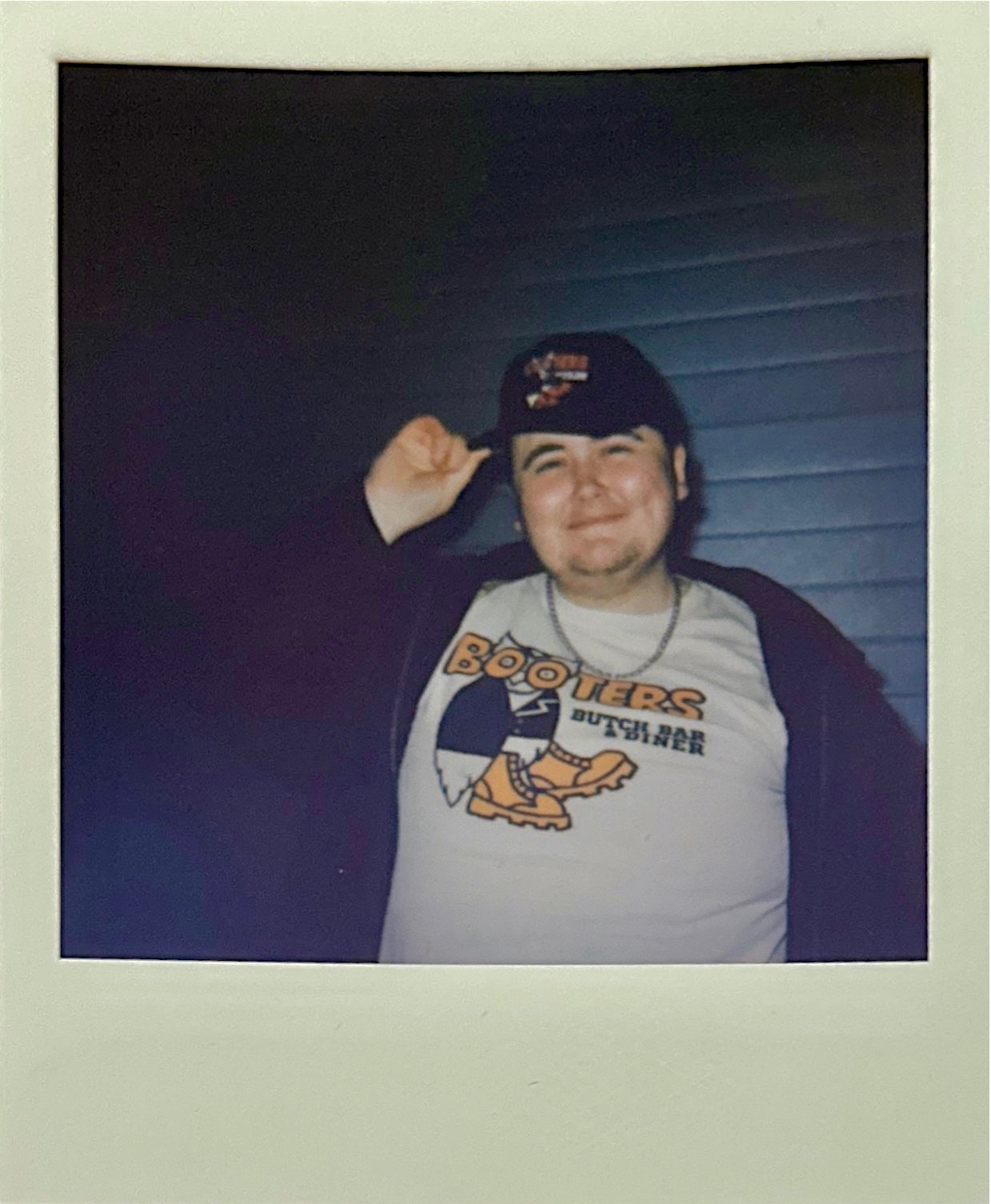 Polaroid of person wearing black cap and white Hooters shirt with cartoon owl logo, standing against blue wooden siding at night.