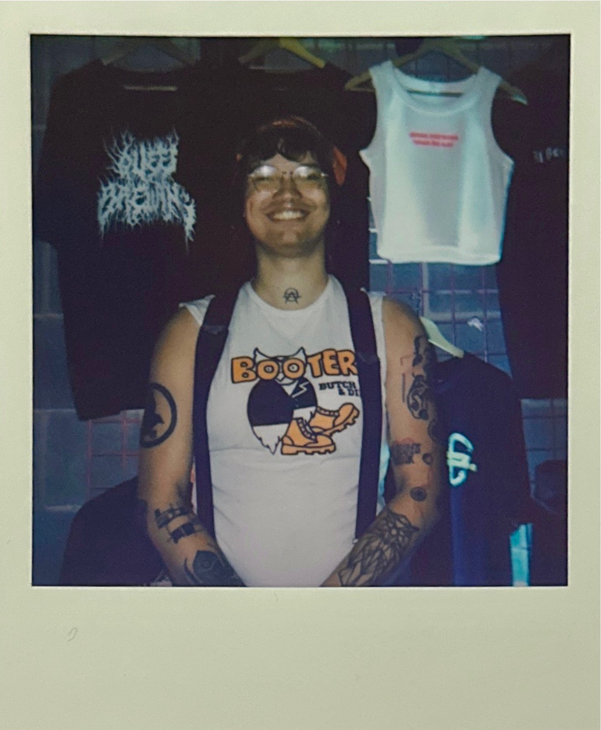 Person with glasses and bandana wearing white vest with cartoon character design, standing in clothing shop with hanging garments.