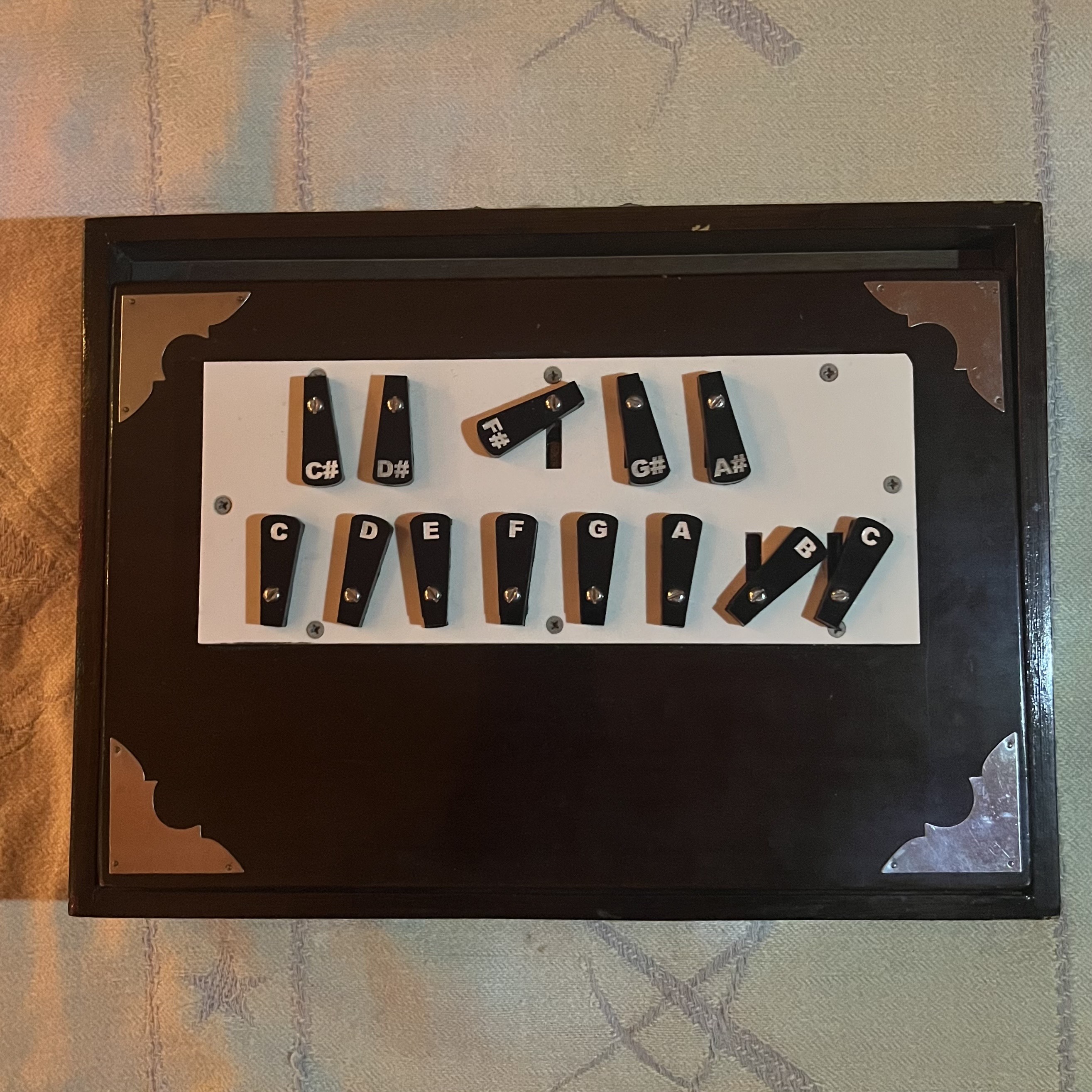 Black wooden frame containing white panel with dark wooden pieces arranged in rows, each piece marked with letters like C#, D#, F#, G#, A#.