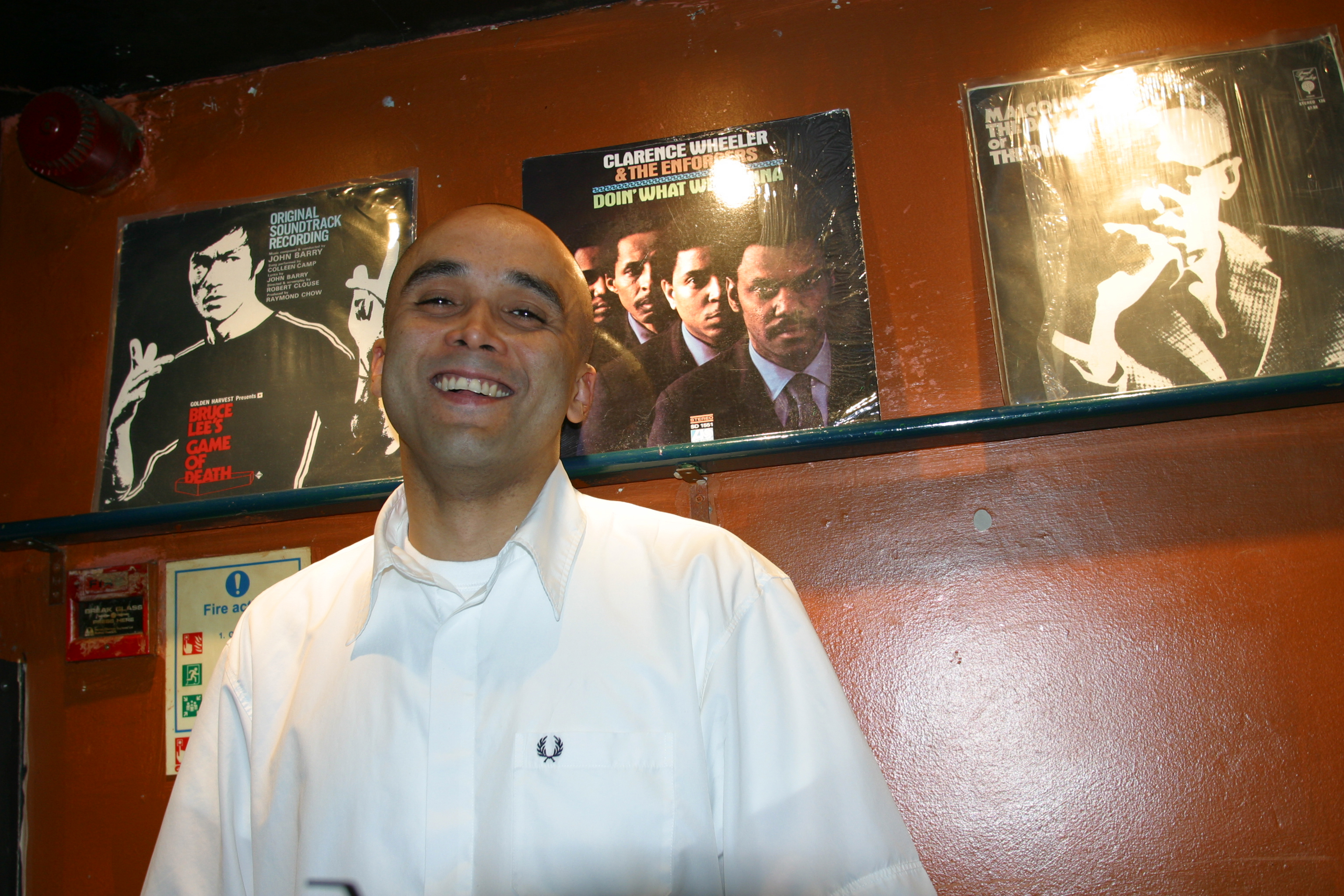 Smiling bald man in white Fred Perry polo shirt standing against orange wall with framed film posters including The Godfather.