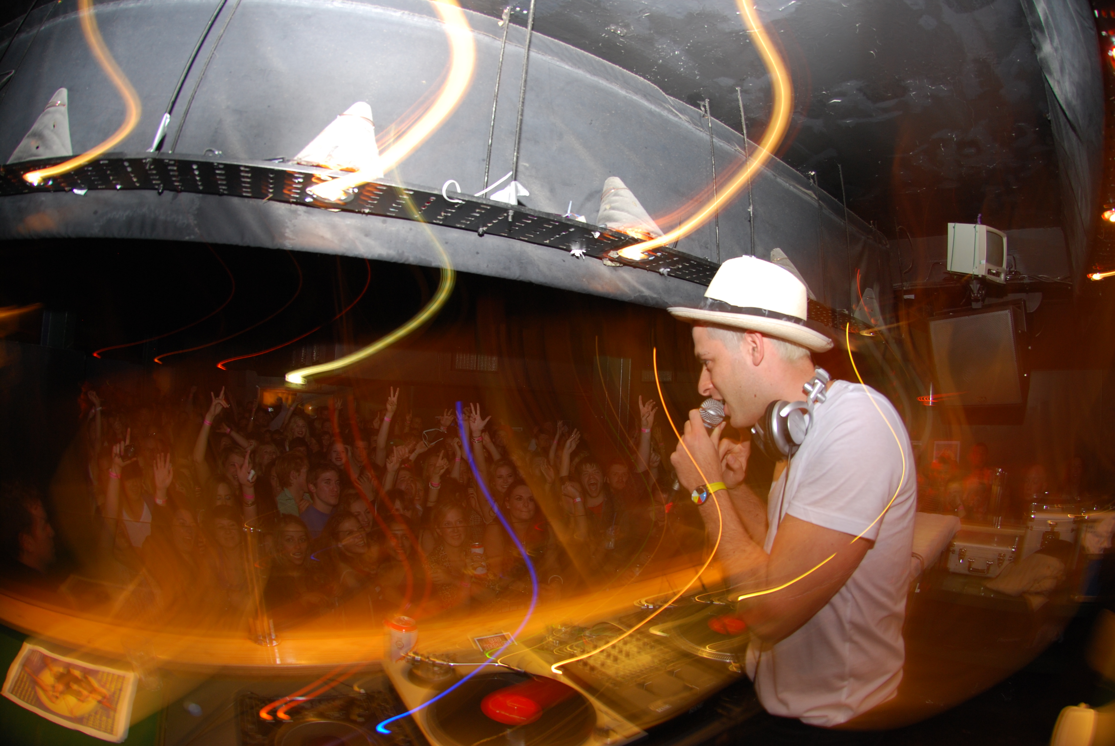 DJ in white shirt and hat at turntables with long-exposure light trails creating yellow and orange streaks across dark venue.