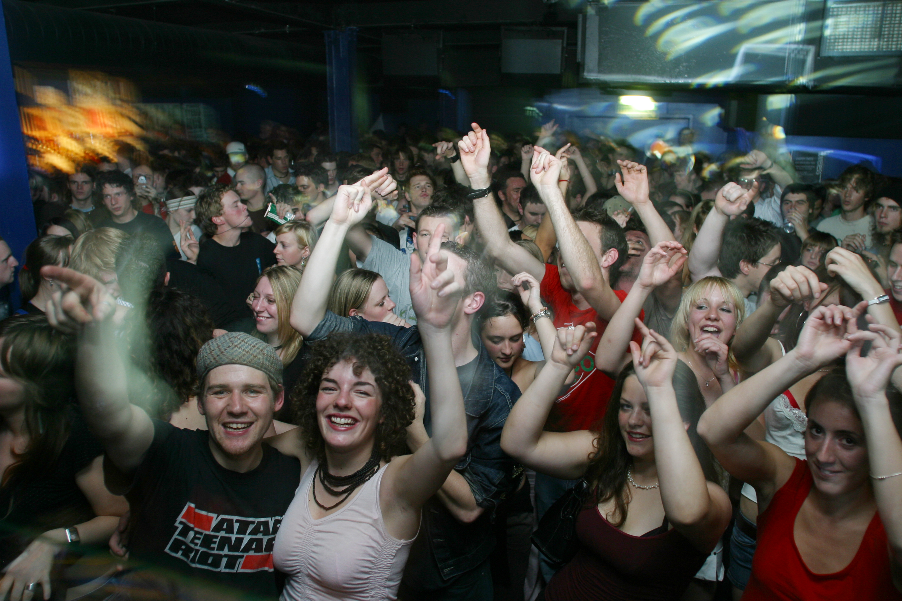 Crowded nightclub scene with people dancing, arms raised, under blue and green lighting with motion blur effects.