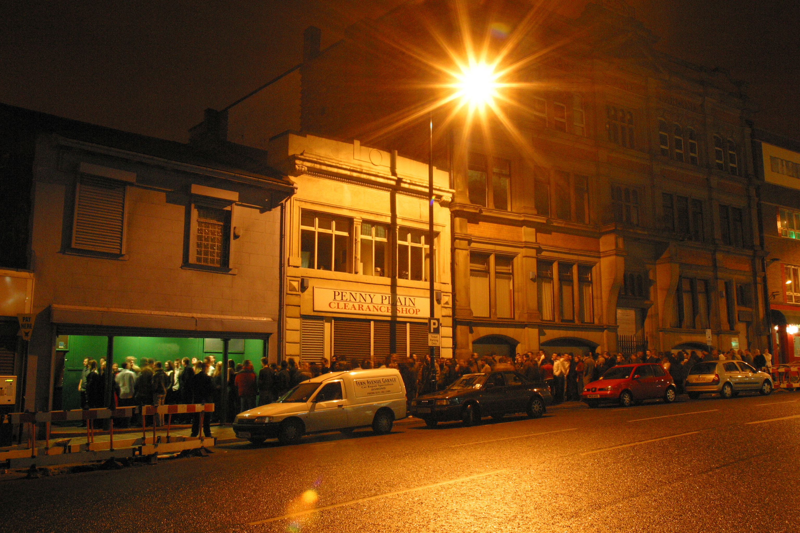 Night street scene with orange streetlight illuminating two-storey buildings, parked cars, and people gathered outside shops.