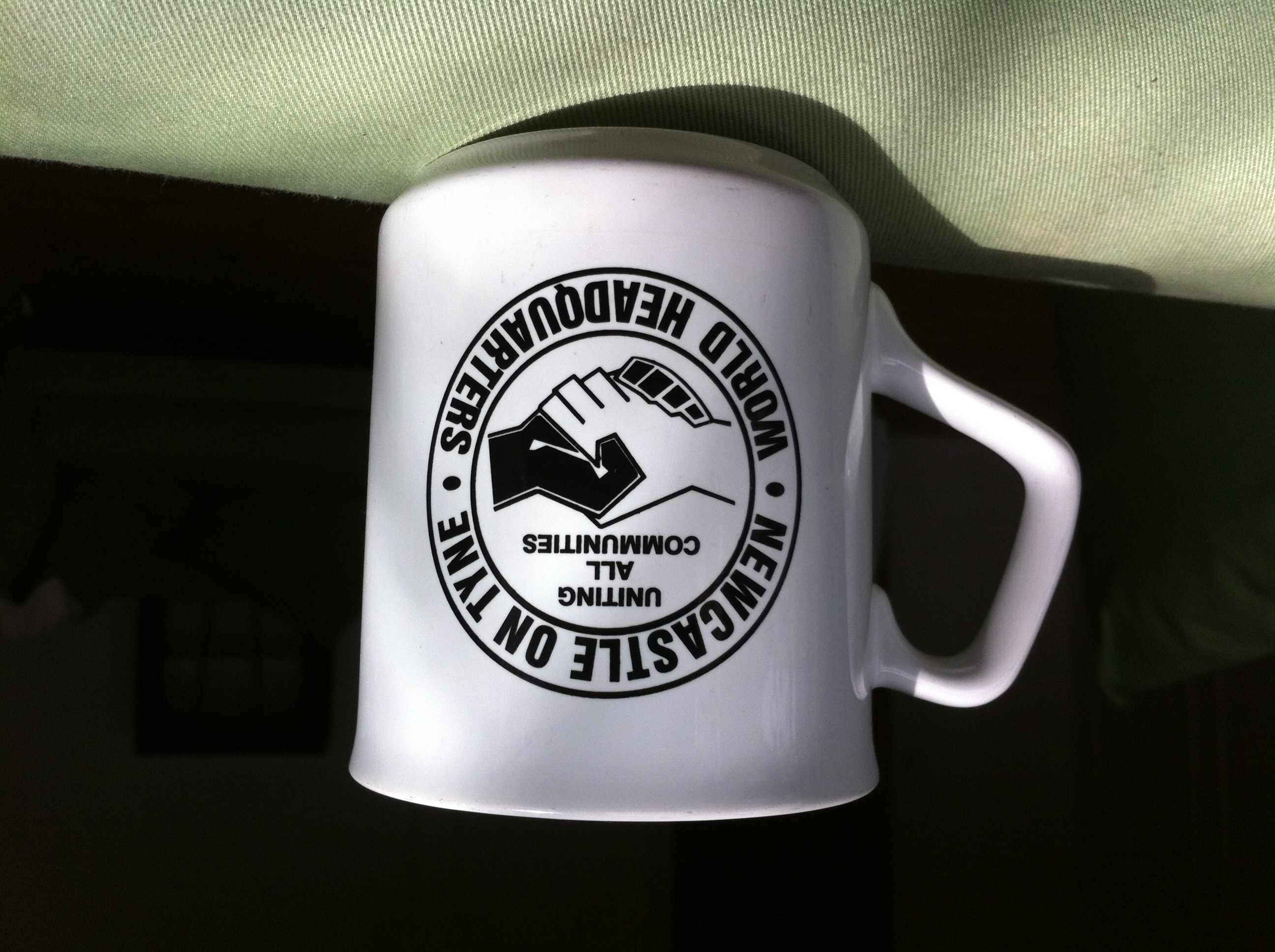 White ceramic mug with black circular logo reading "WORLD HEADQUARTERS UNITED ALL COMMUNITIES" around mountain and handshake design.