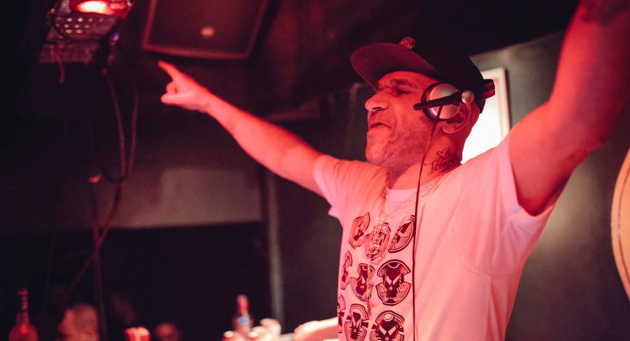 DJ with glasses and white t-shirt raising both arms whilst performing, bathed in red stage lighting in dark venue.