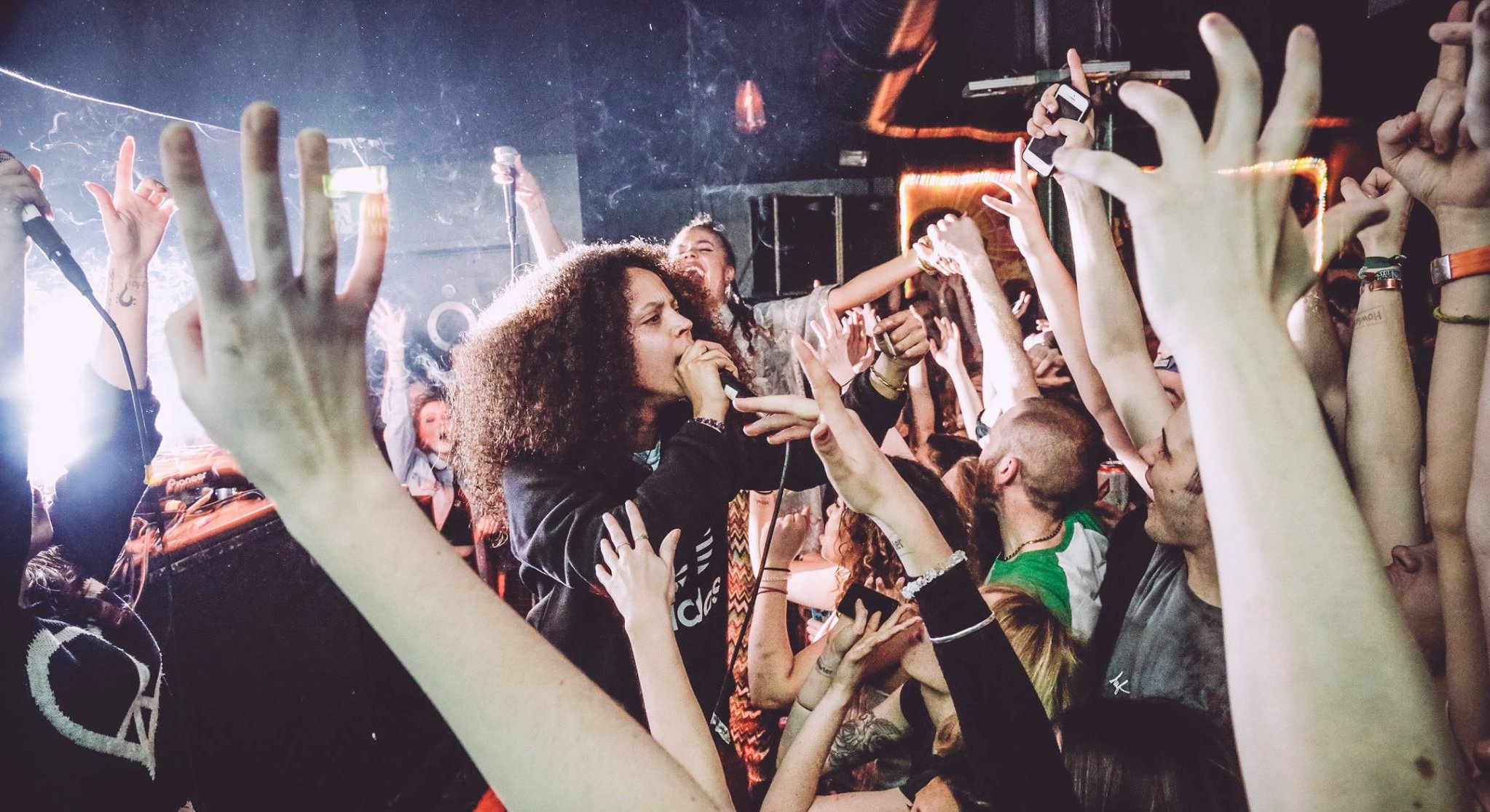 Performer with curly hair singing into microphone surrounded by numerous raised hands reaching upward in dimly lit venue.