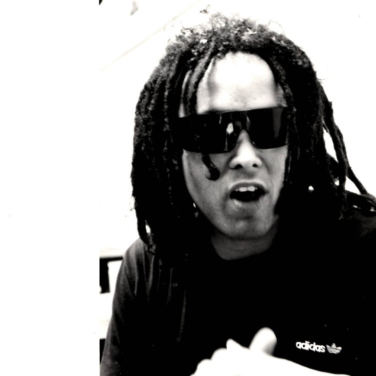 Black and white image of person with dreadlocks wearing dark sunglasses and Adidas tracksuit top, mouth open, gesturing with hand.