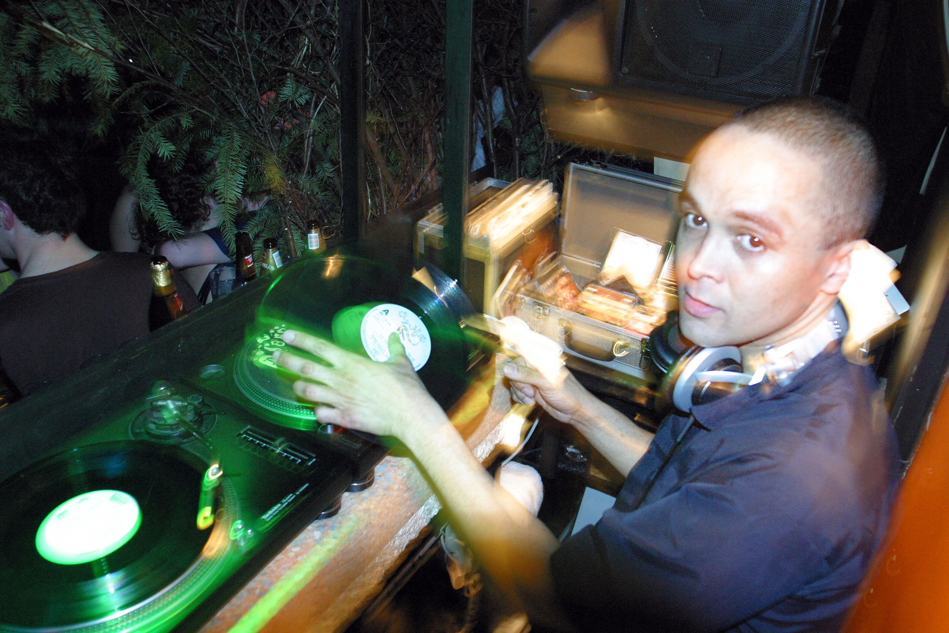 DJ with headphones operating green-lit turntables in dimly lit booth, motion blur on hands from mixing records.