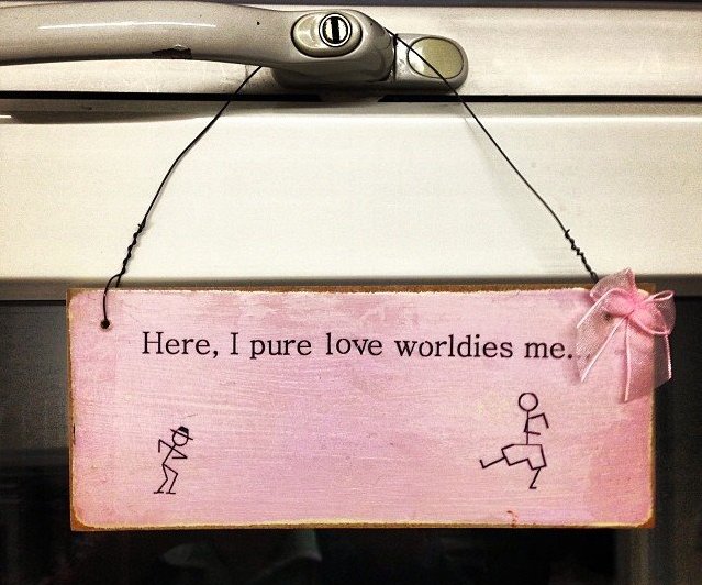 Pink wooden sign hanging from beige door handle with black text "Here, I pure love worldies me" and simple stick figure drawings.