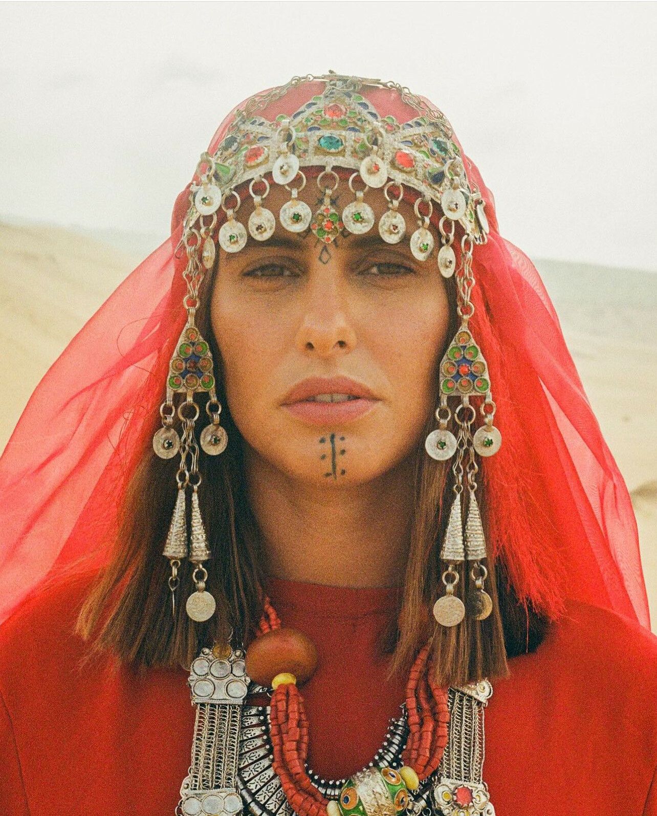 Ornate headpiece with colourful metallic discs, beads and fabric, worn by a person with a serious expression in an arid landscape.