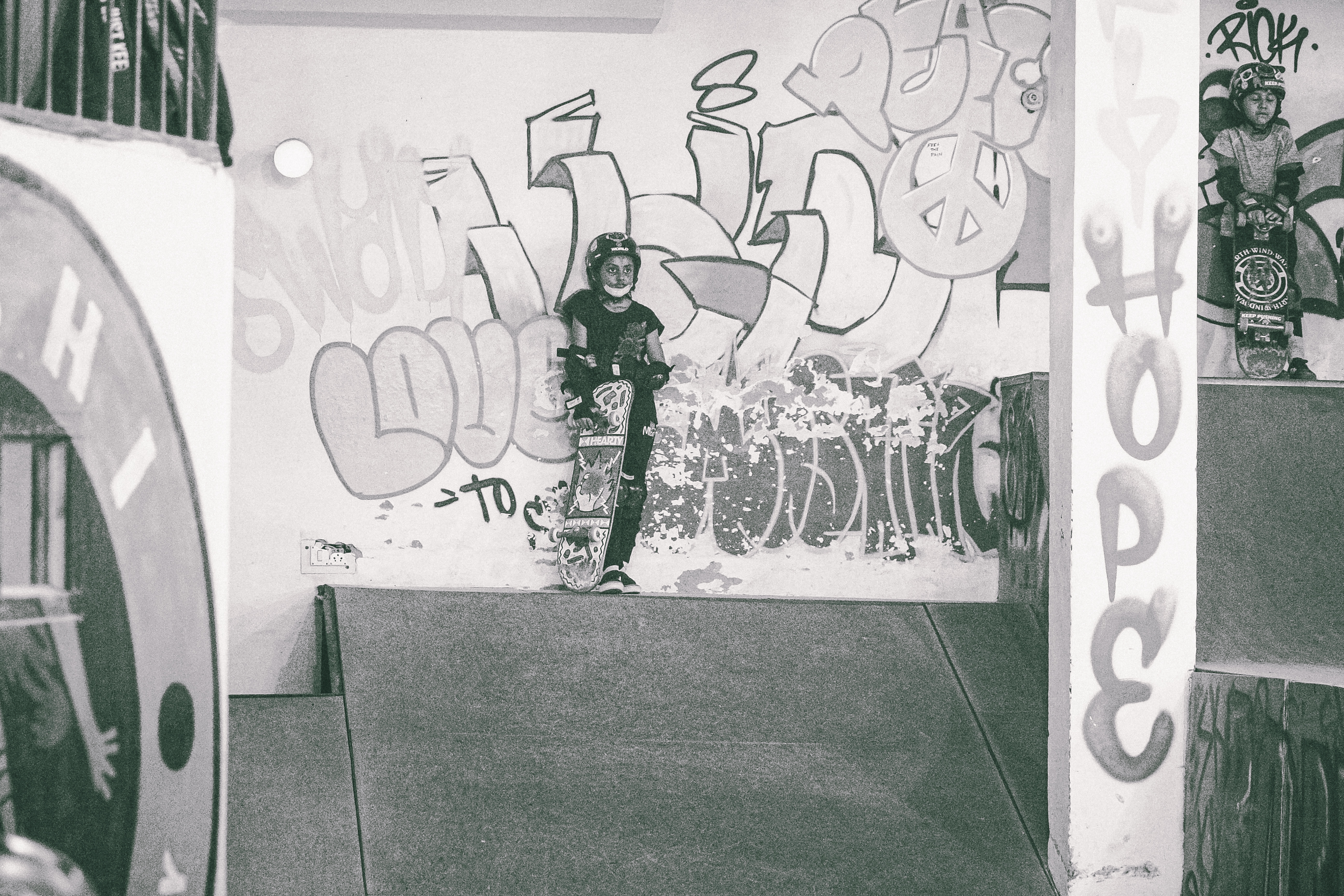 Person in dark clothing standing in concrete underpass with white walls covered in black and pink graffiti lettering and tags.