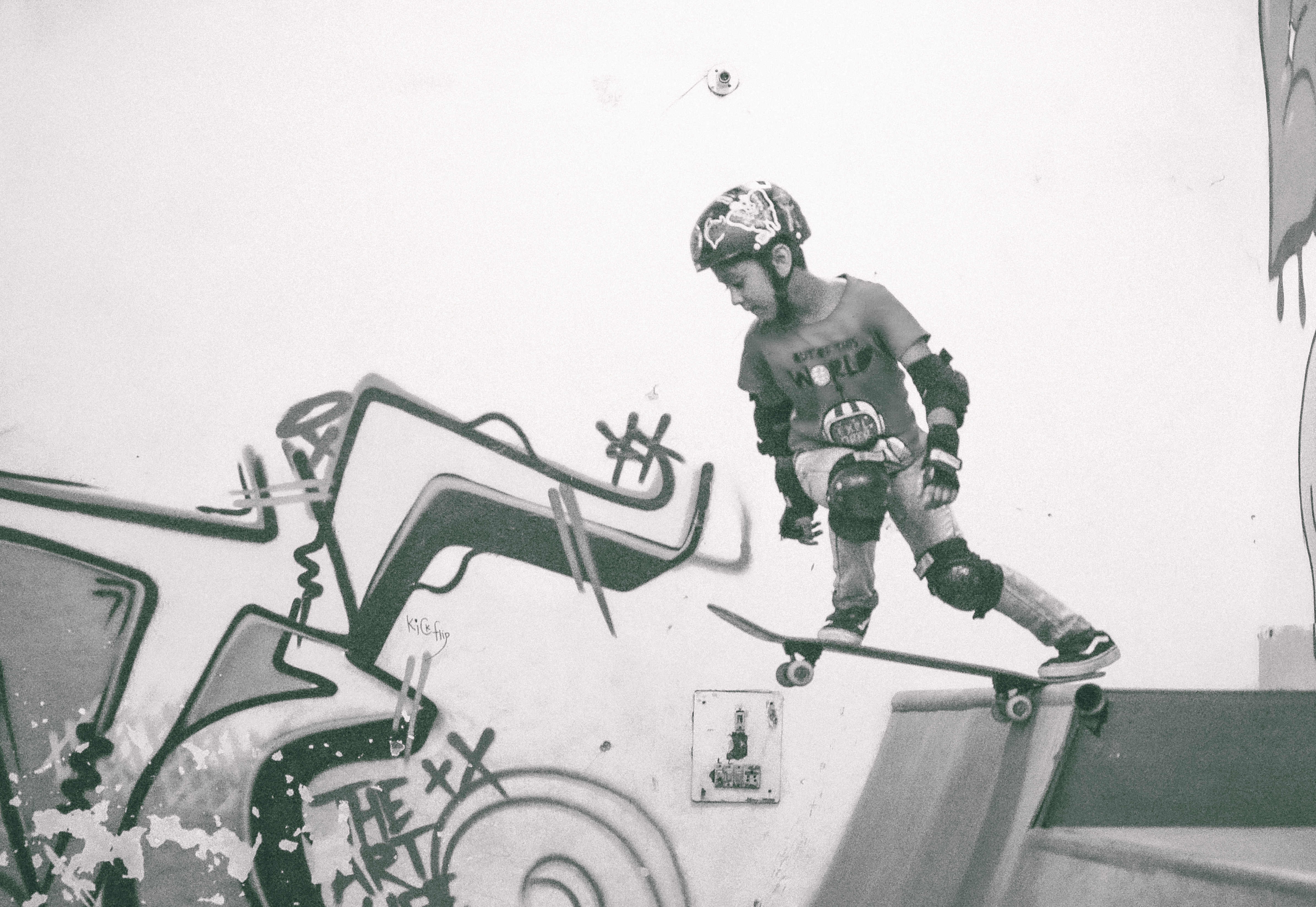 Skateboarder in protective gear on ramp with green and black graffiti on white wall, grey and green colour scheme.