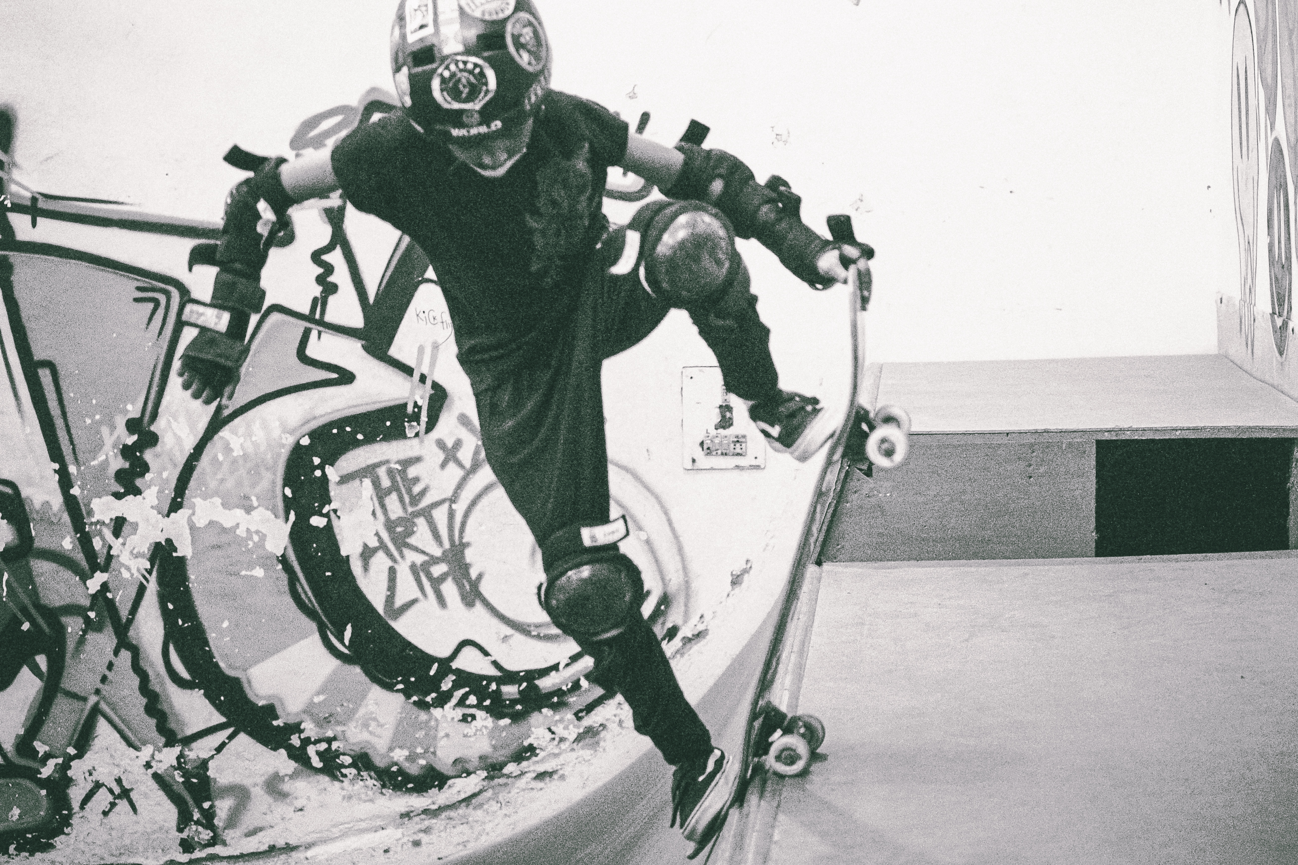 Person in protective gear riding white bicycle with large circular frame in concrete skate bowl, black and white image.