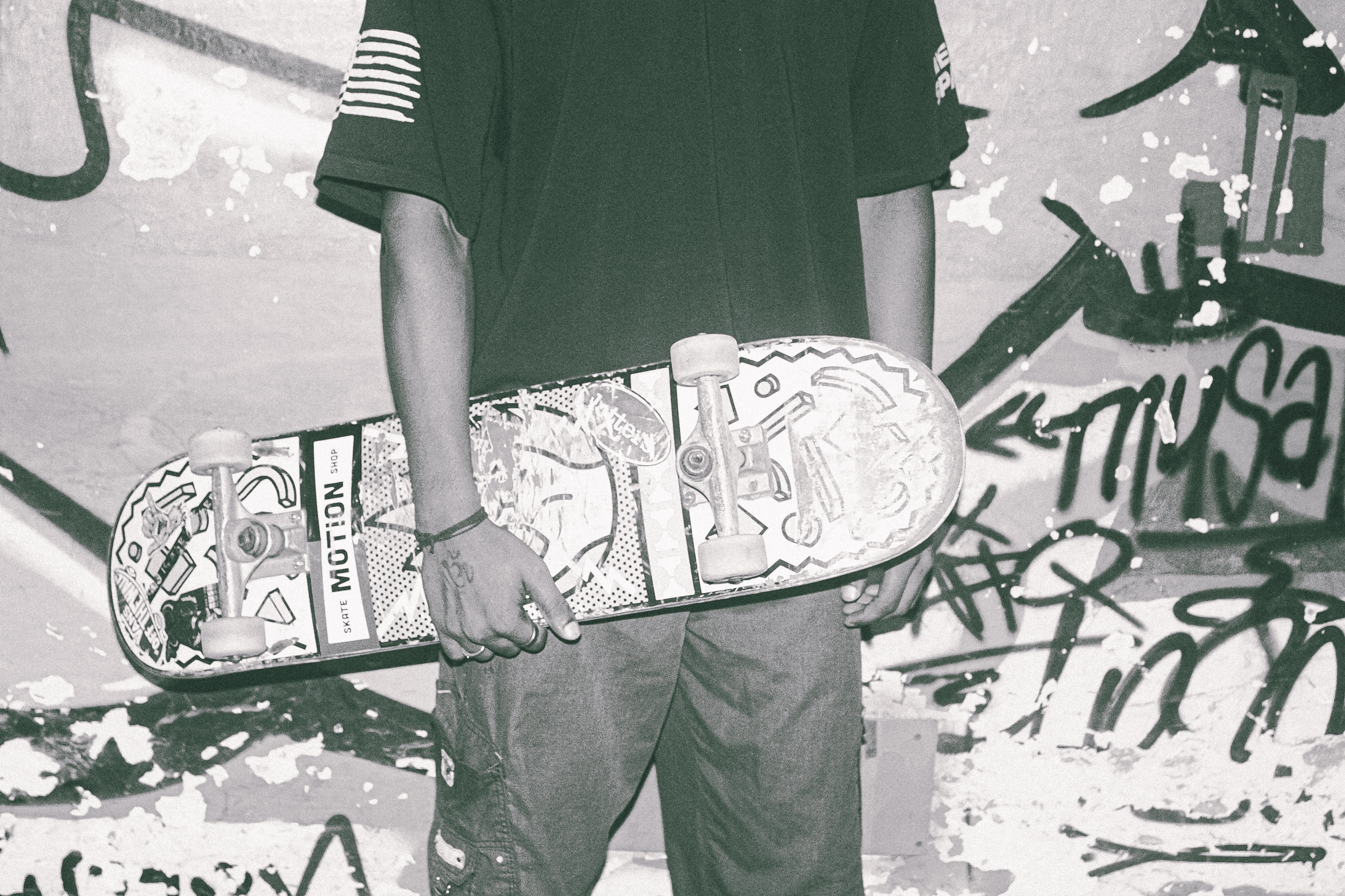 Person in dark green top holding decorated skateboard, standing against wall covered in black graffiti and markings.