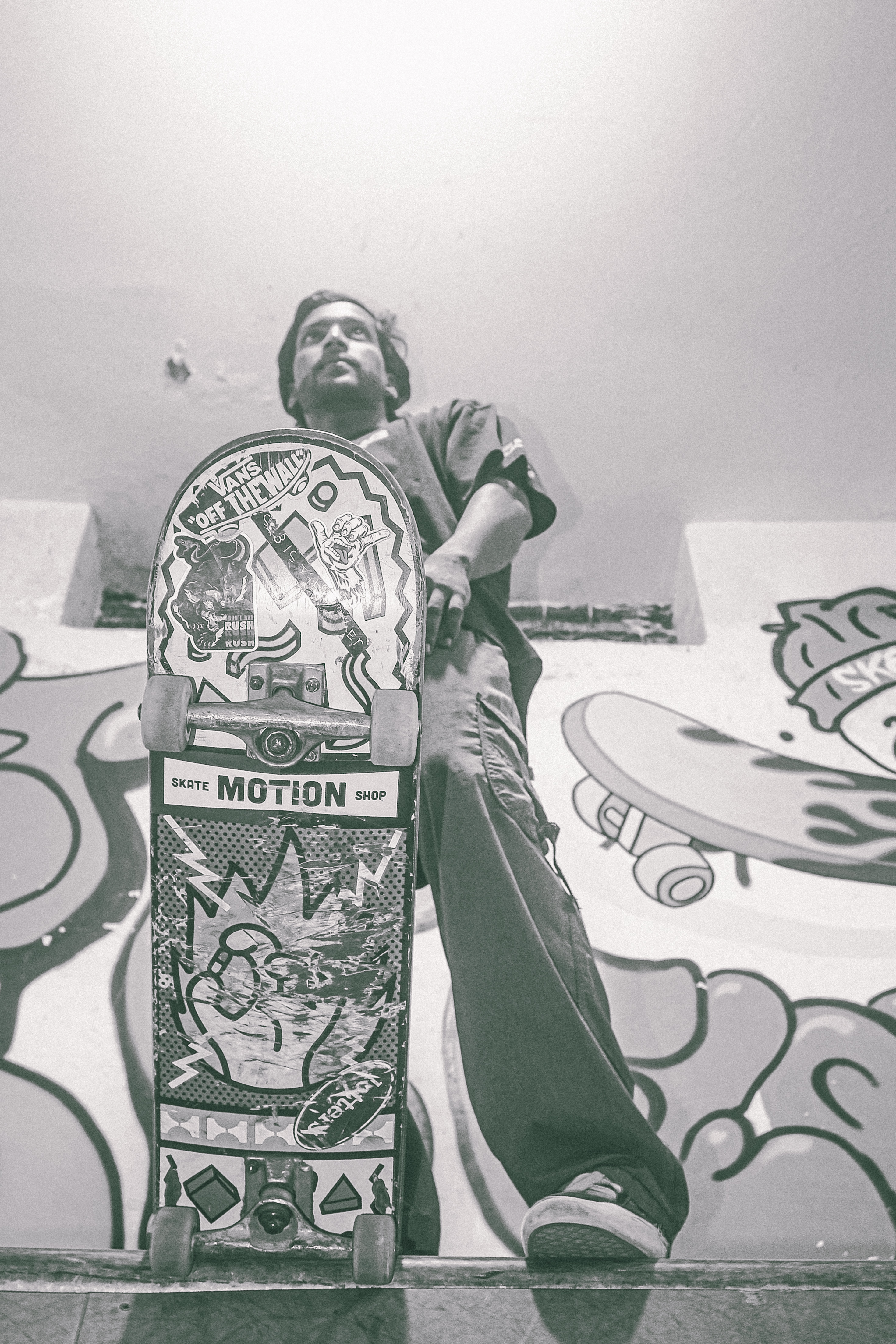 Man with beard holding decorated skateboard against graffitied wall, black and white image with pink-tinted sky background.