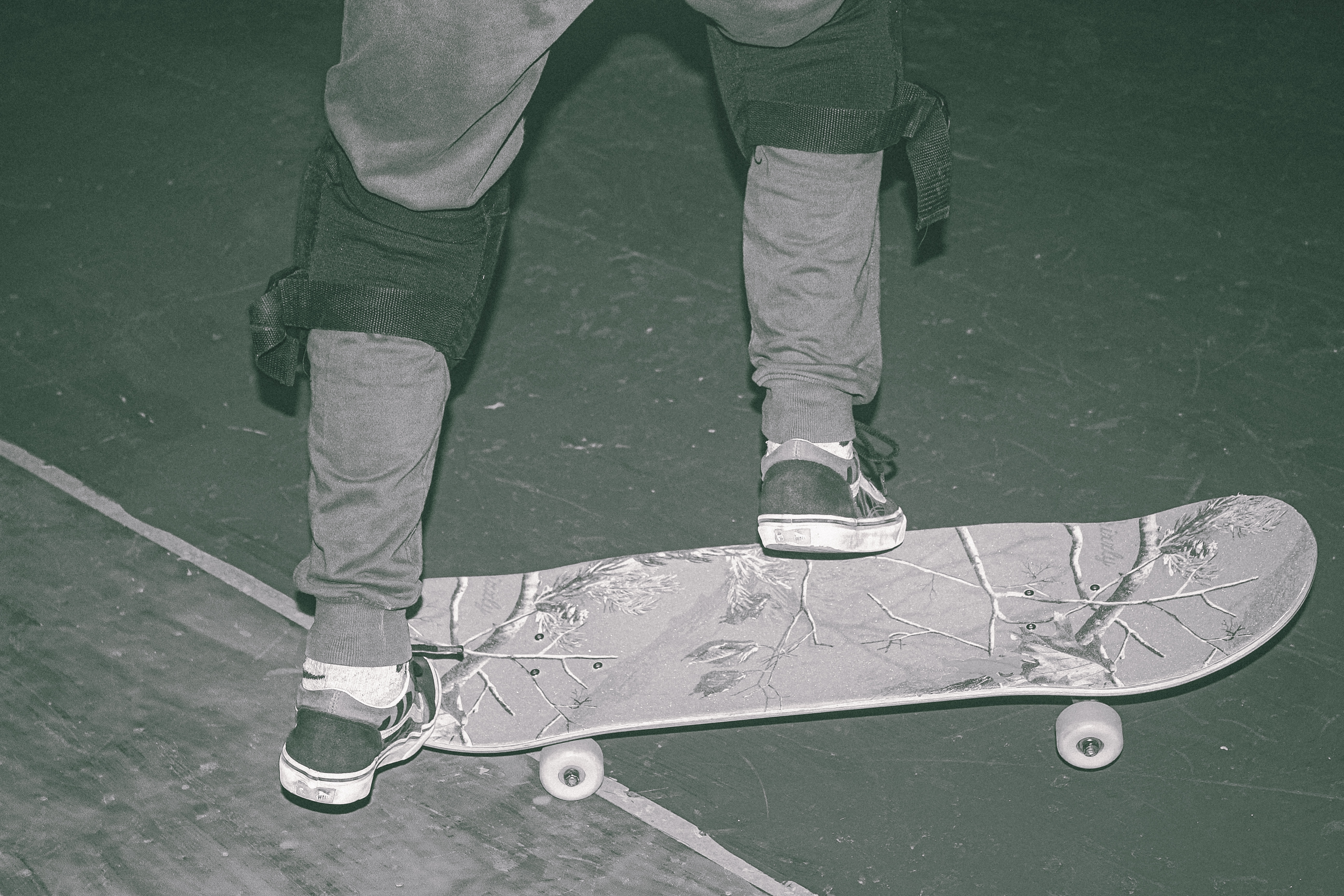 Person's legs and feet on weathered skateboard with white wheels on green tarmac surface.