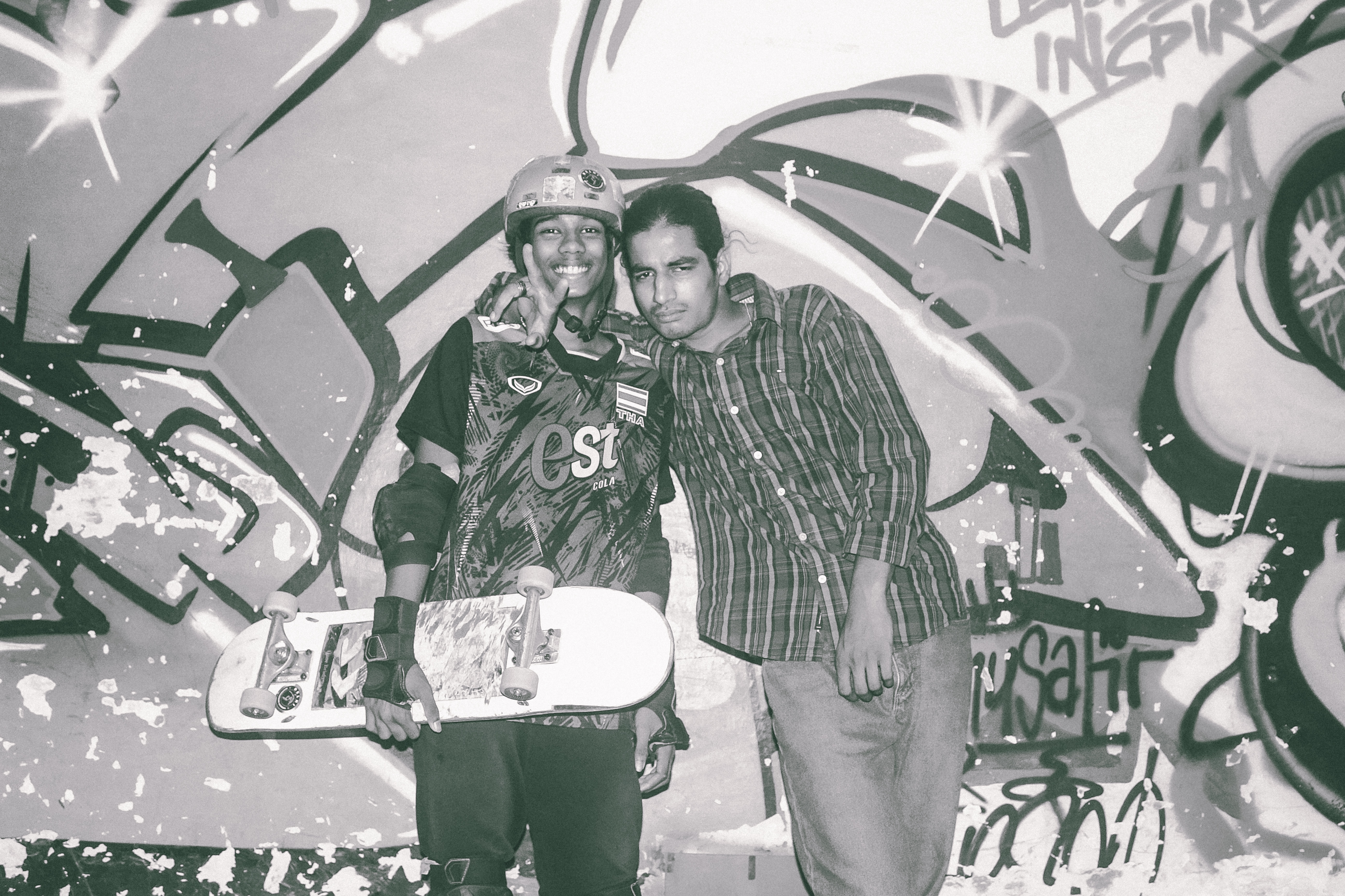 Two men posing together in front of colourful graffiti wall with circular and curved designs in green, white, and grey tones.