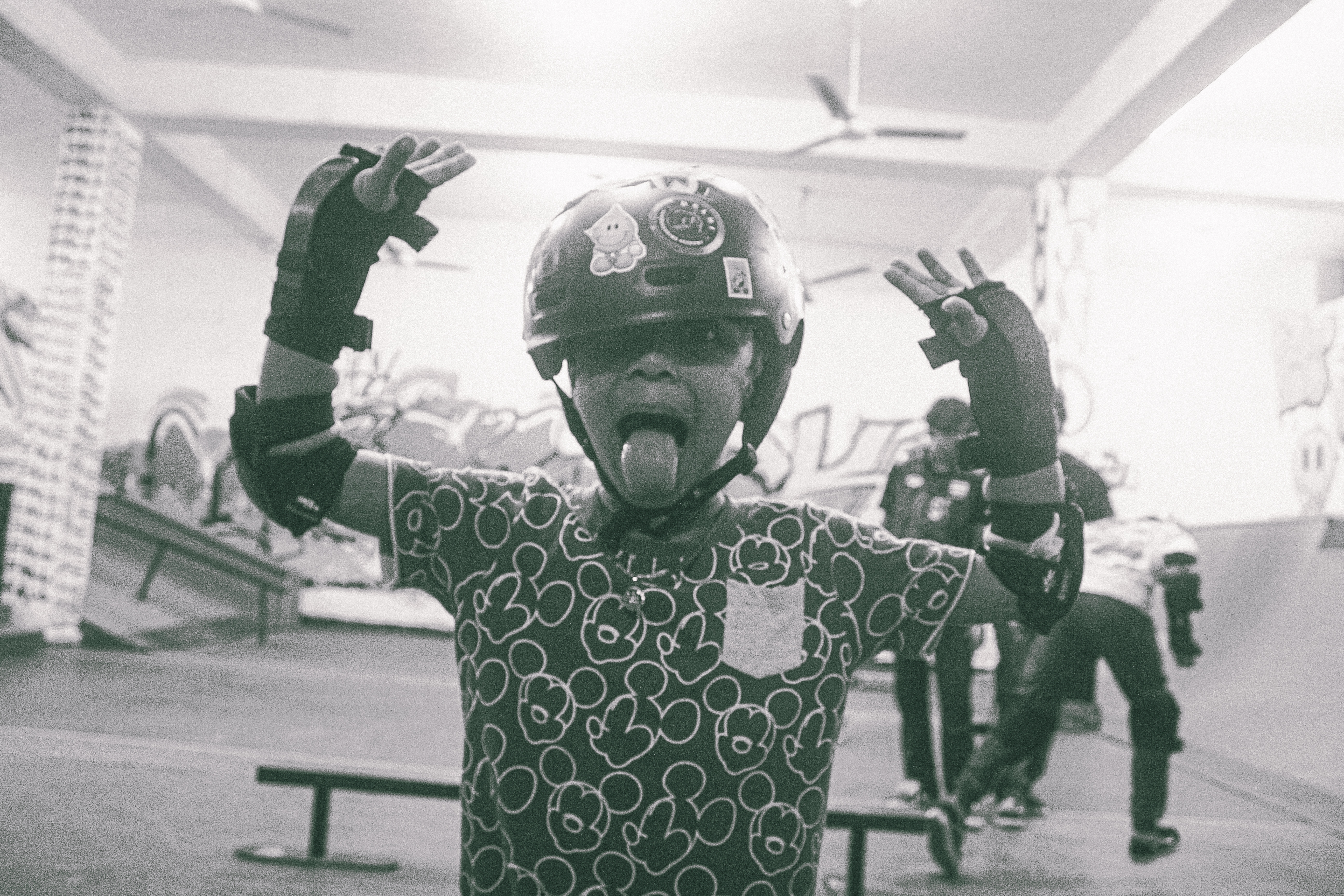 Child in helmet and patterned shirt with arms raised, sitting on bench in skate park with ramps visible in background.