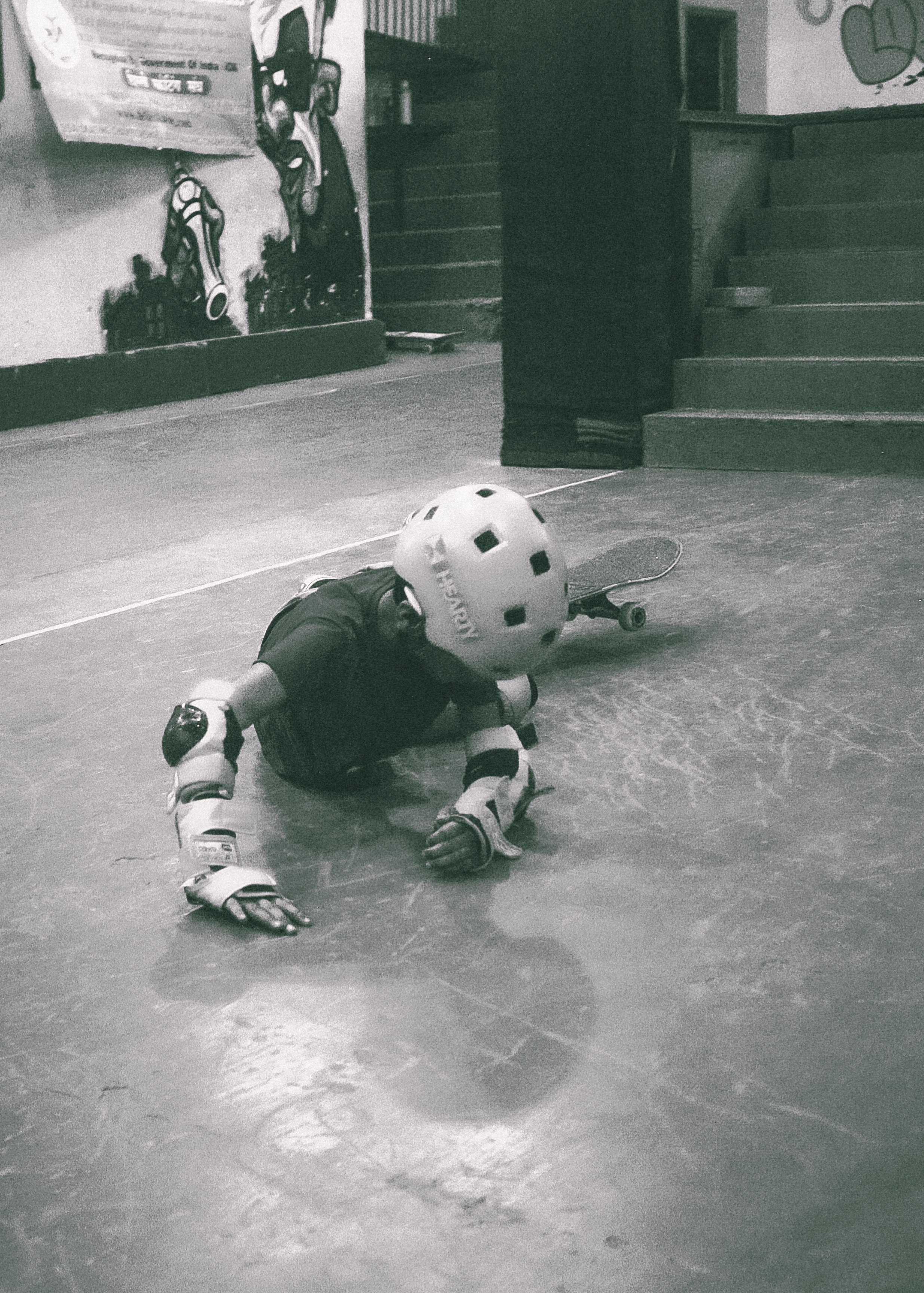Person in protective helmet and knee pads crawling on concrete floor near stairs in what appears to be a training facility.