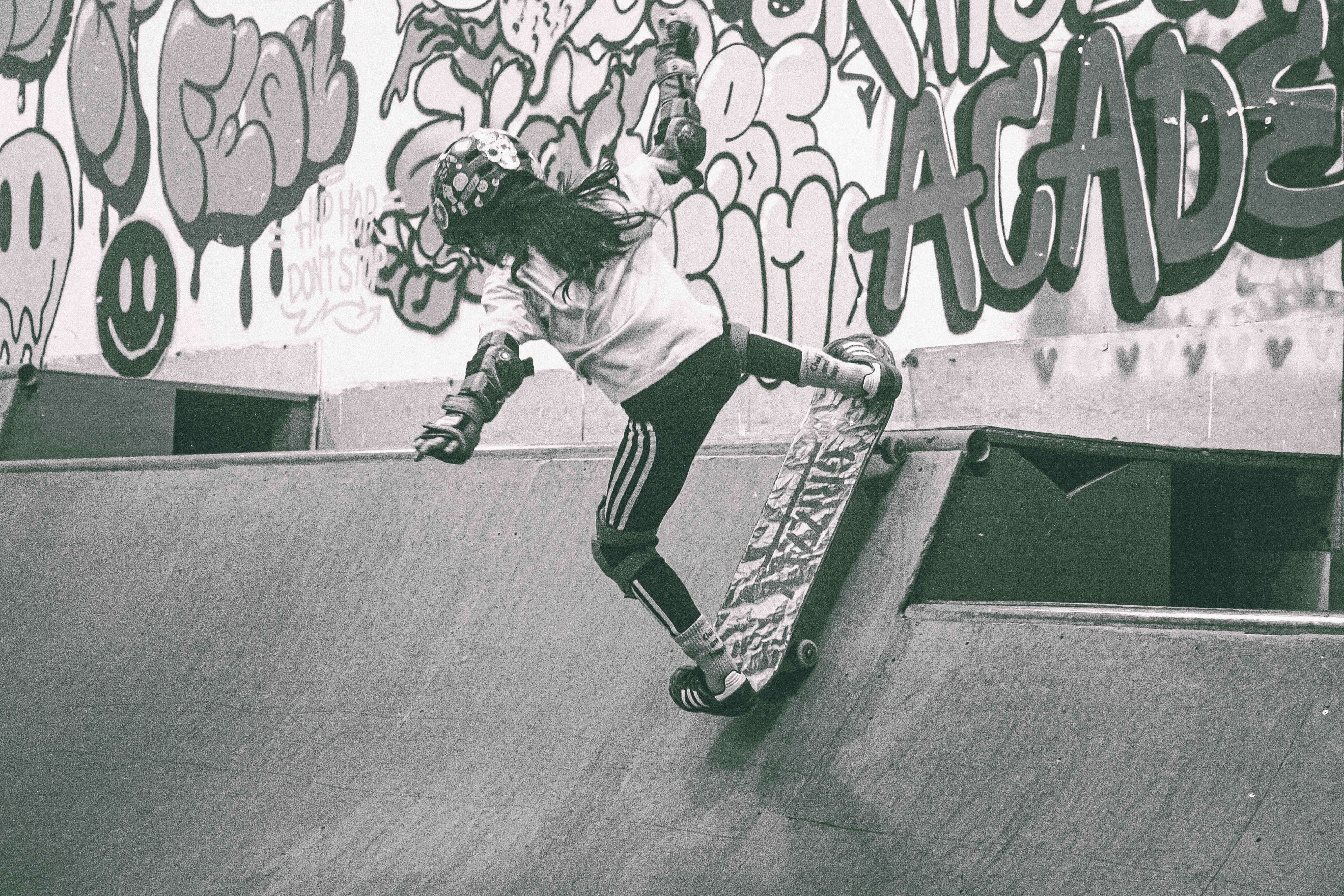 Skateboarder in mid-air above concrete bowl with graffitied wall behind, monochrome tones with green accents on clothing and text.