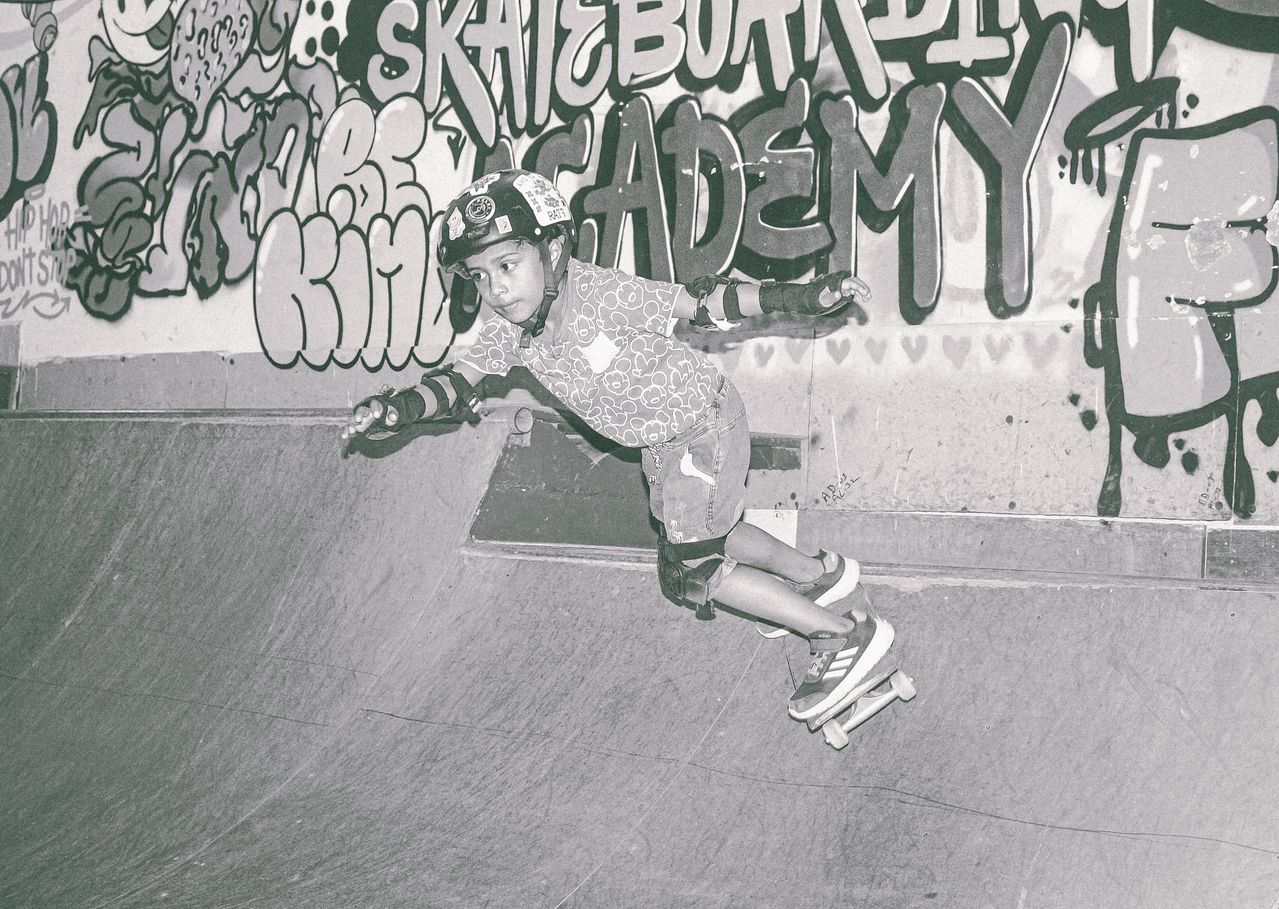 Skateboarder mid-air in bowl with graffiti-covered walls, black and white image with skateboard visible below feet.
