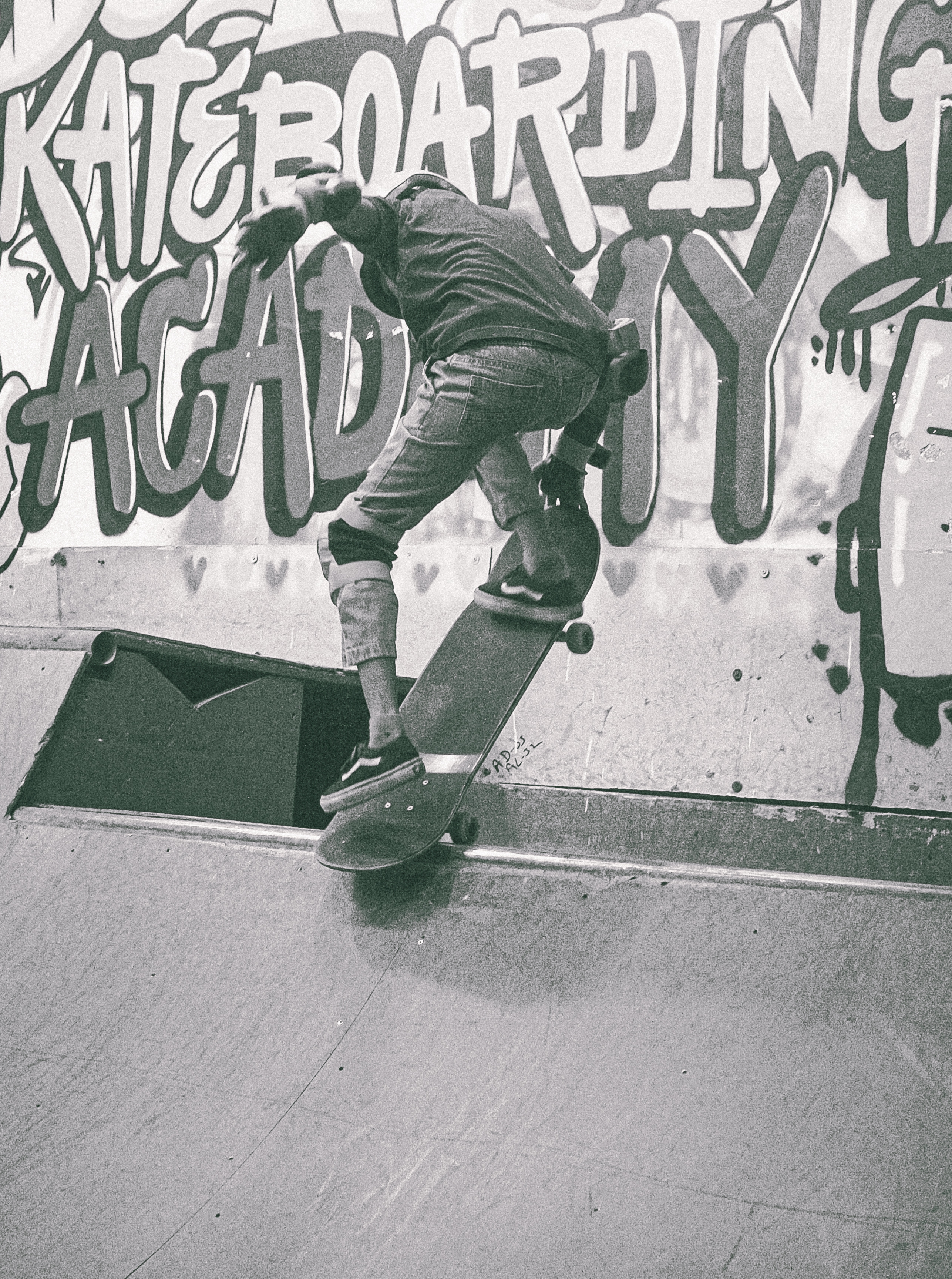 Person on skateboard at concrete skate ramp with white graffiti text on wall behind, black and white tones with slight green tint.
