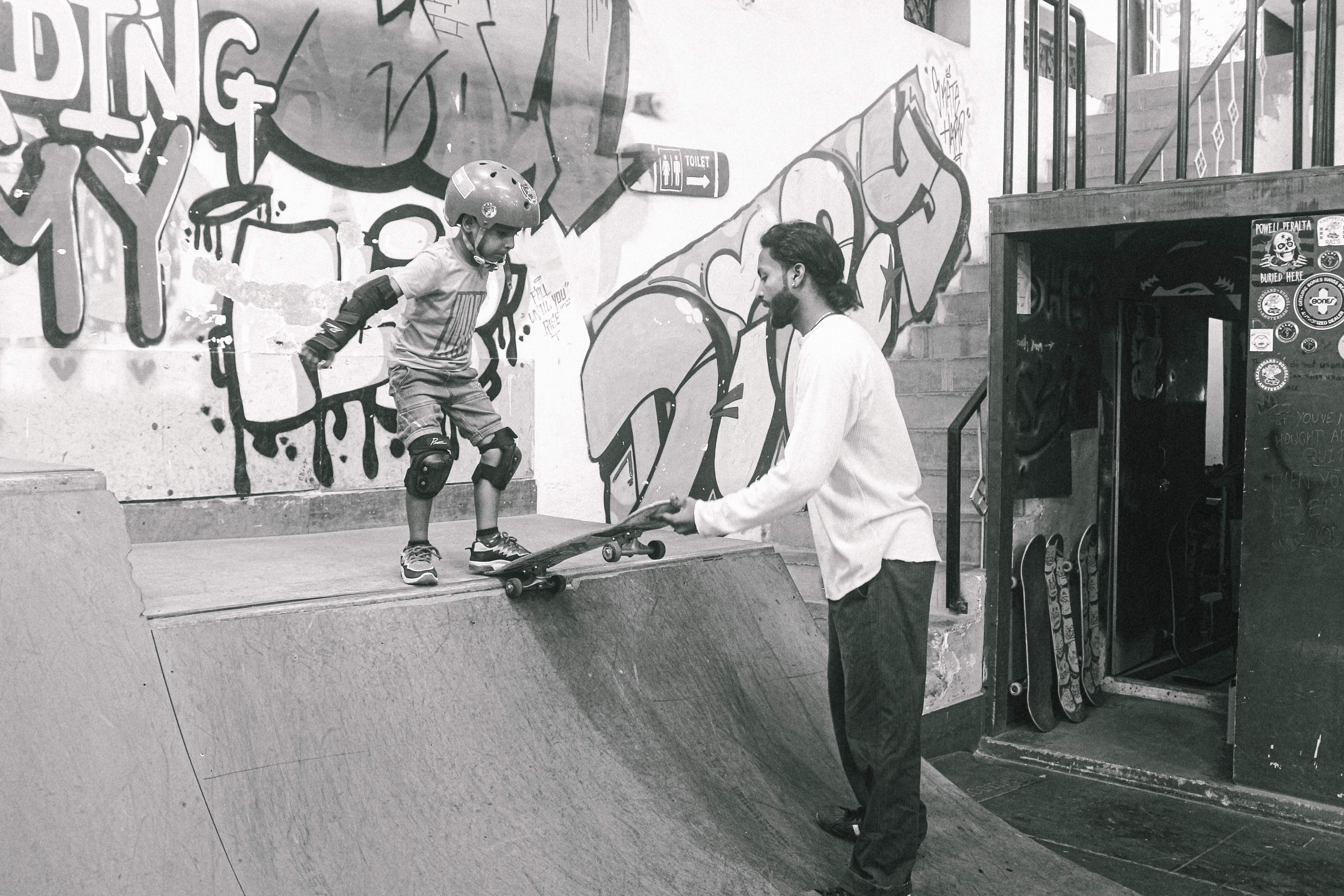 Two skateboarders at concrete skate bowl with graffitied walls, one skating whilst other sits on edge holding skateboard