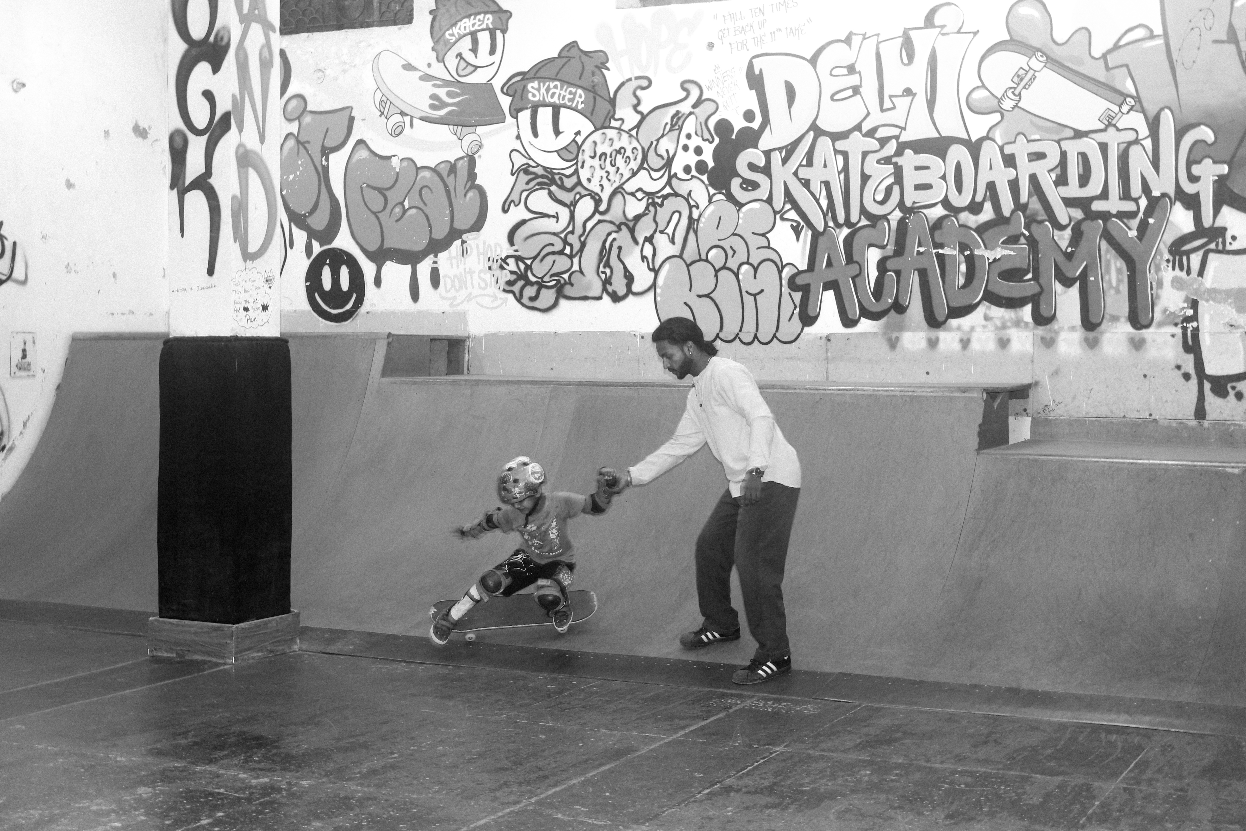 Black and white image of adult helping child skateboard in skate bowl beneath colourful graffiti wall featuring cartoon characters and text.