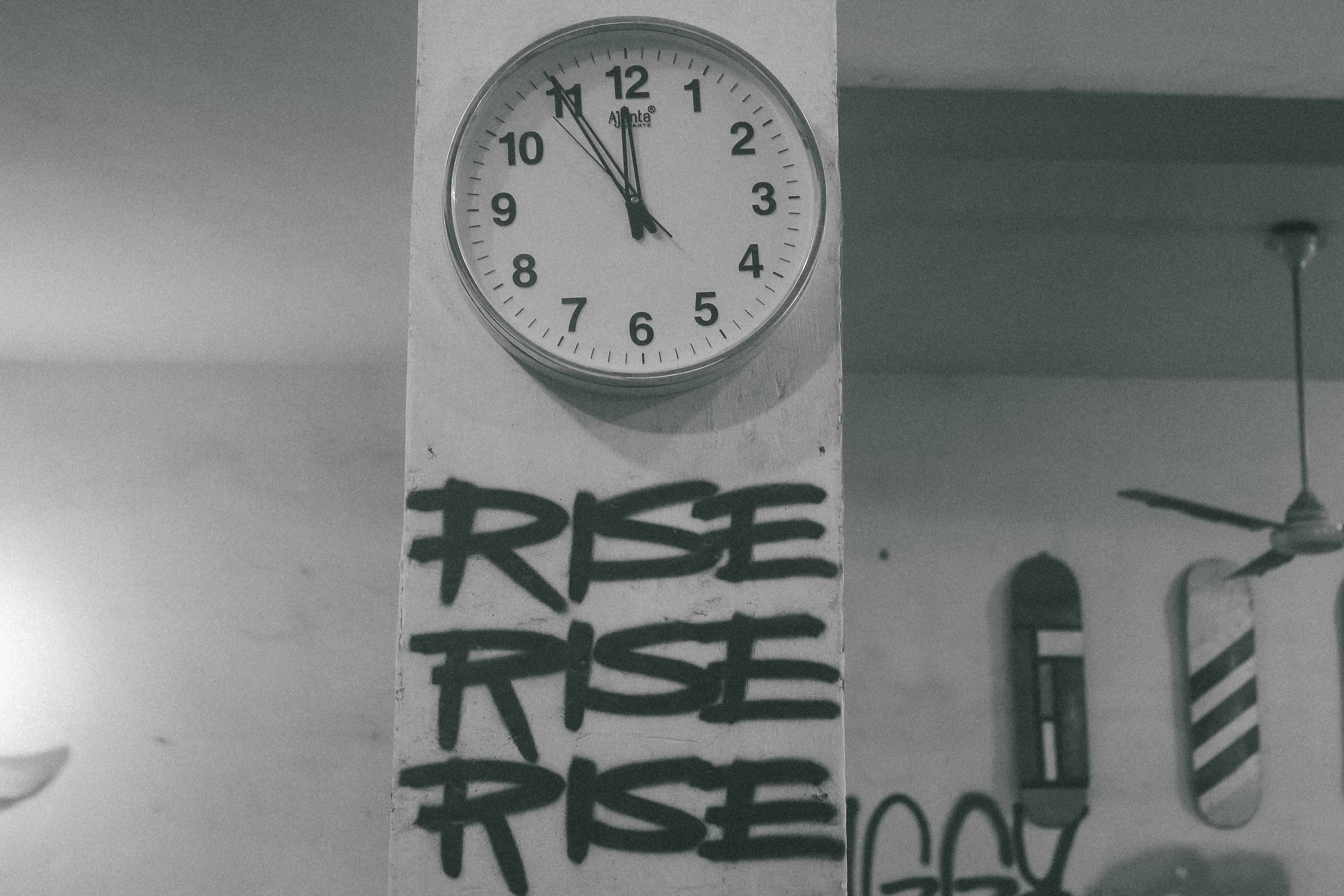 White wall clock showing approximately 11:55, mounted above green graffiti text reading "RISE RISE RISE" in bold lettering.