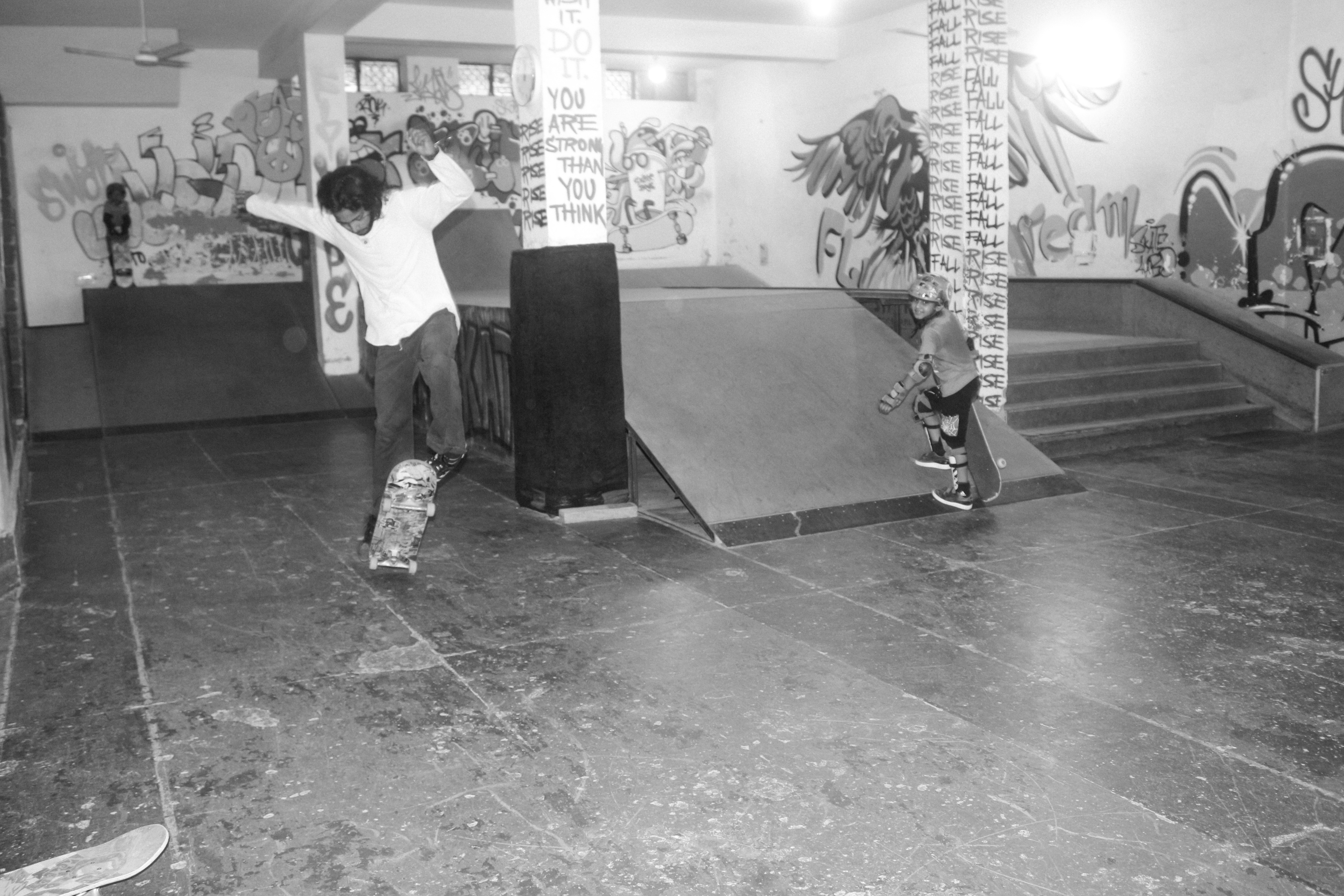 Black and white image of skateboarders at indoor skate park with ramps, graffitied walls, and concrete floor.