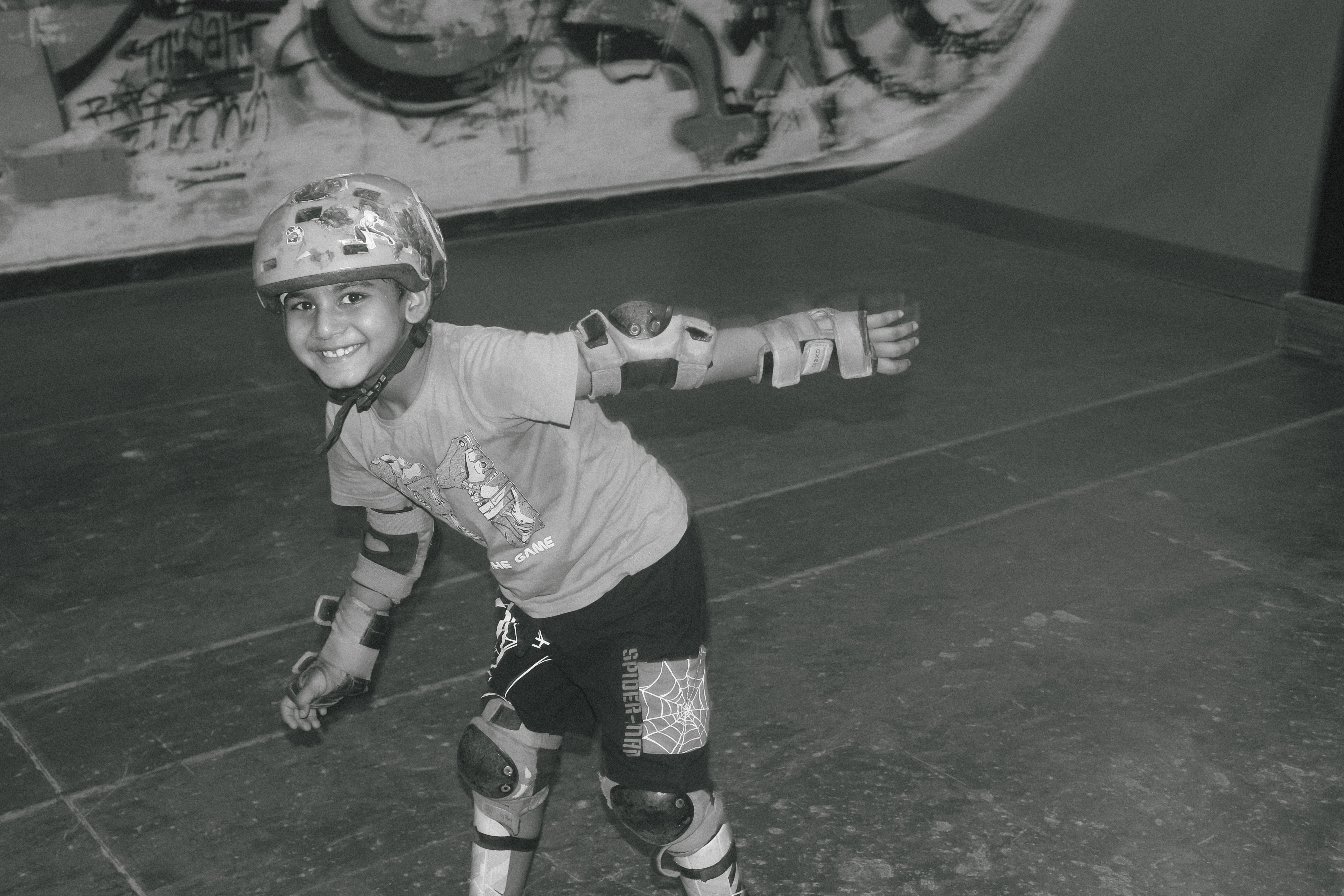 Black and white image of young child in protective helmet and pads, crouched in skating position at skate park with graffiti walls.