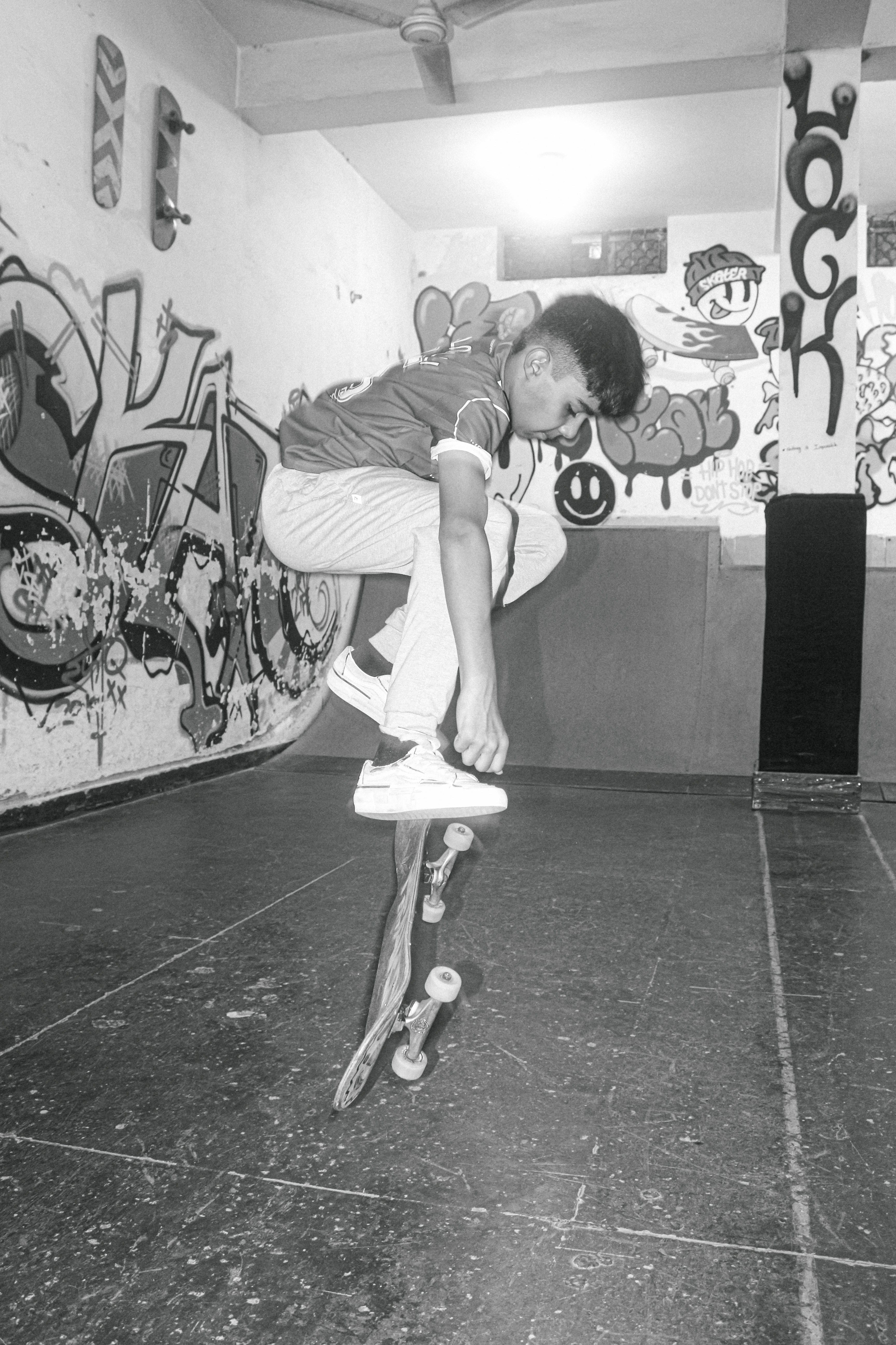 Man performing skateboard trick indoors, graffiti-covered walls, black and white image, ceiling fan visible above.