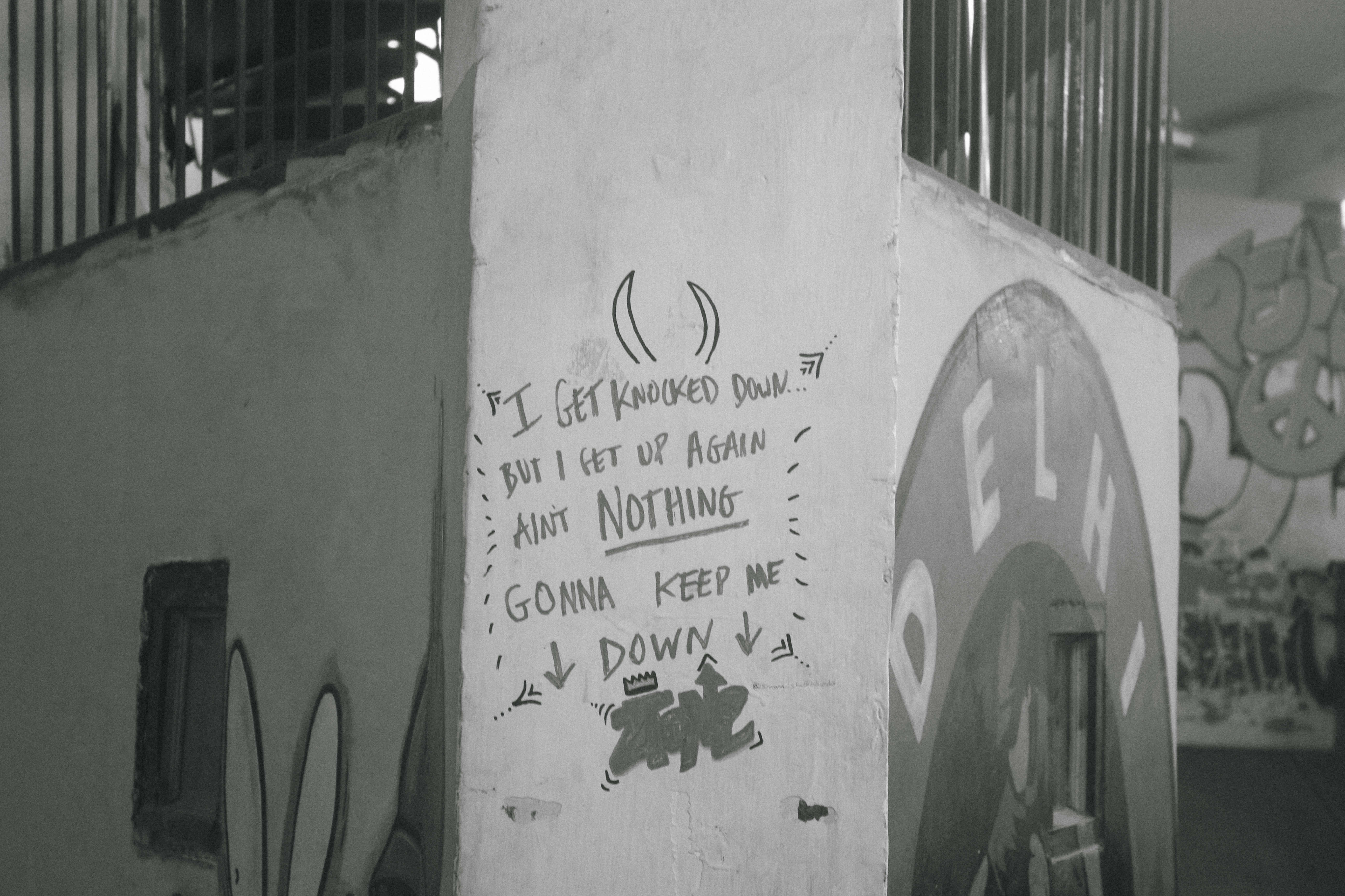 Handwritten text on white pillar reading "I get knocked down but I get up again ain't nothing gonna keep me down" in black and white image.
