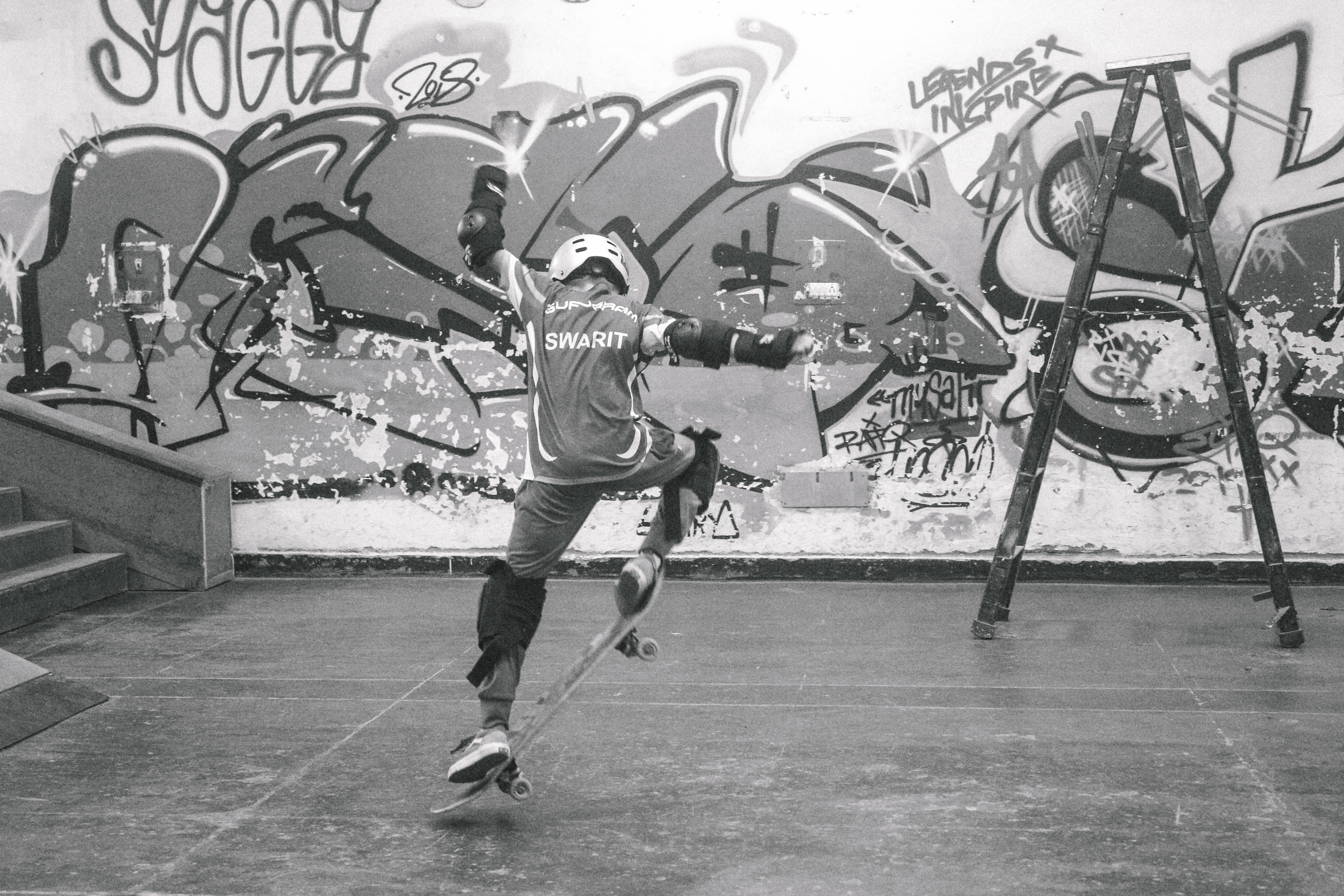 Person skateboarding on concrete surface with large graffiti-covered wall behind them, black and white image with playground equipment visible.