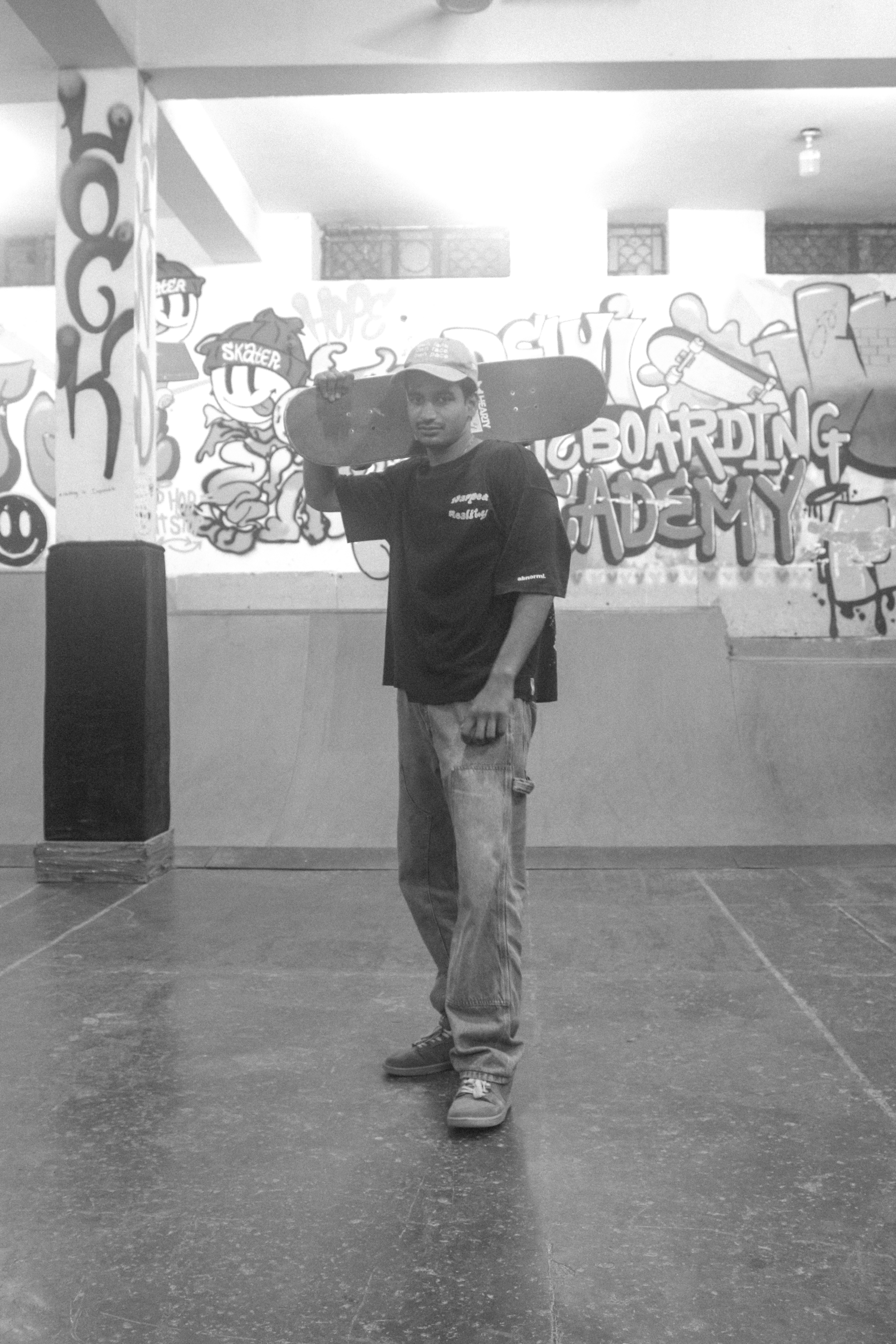 Person holding skateboard in indoor space with graffiti-covered walls, black and white image with tiled floor and exposed ceiling beams.