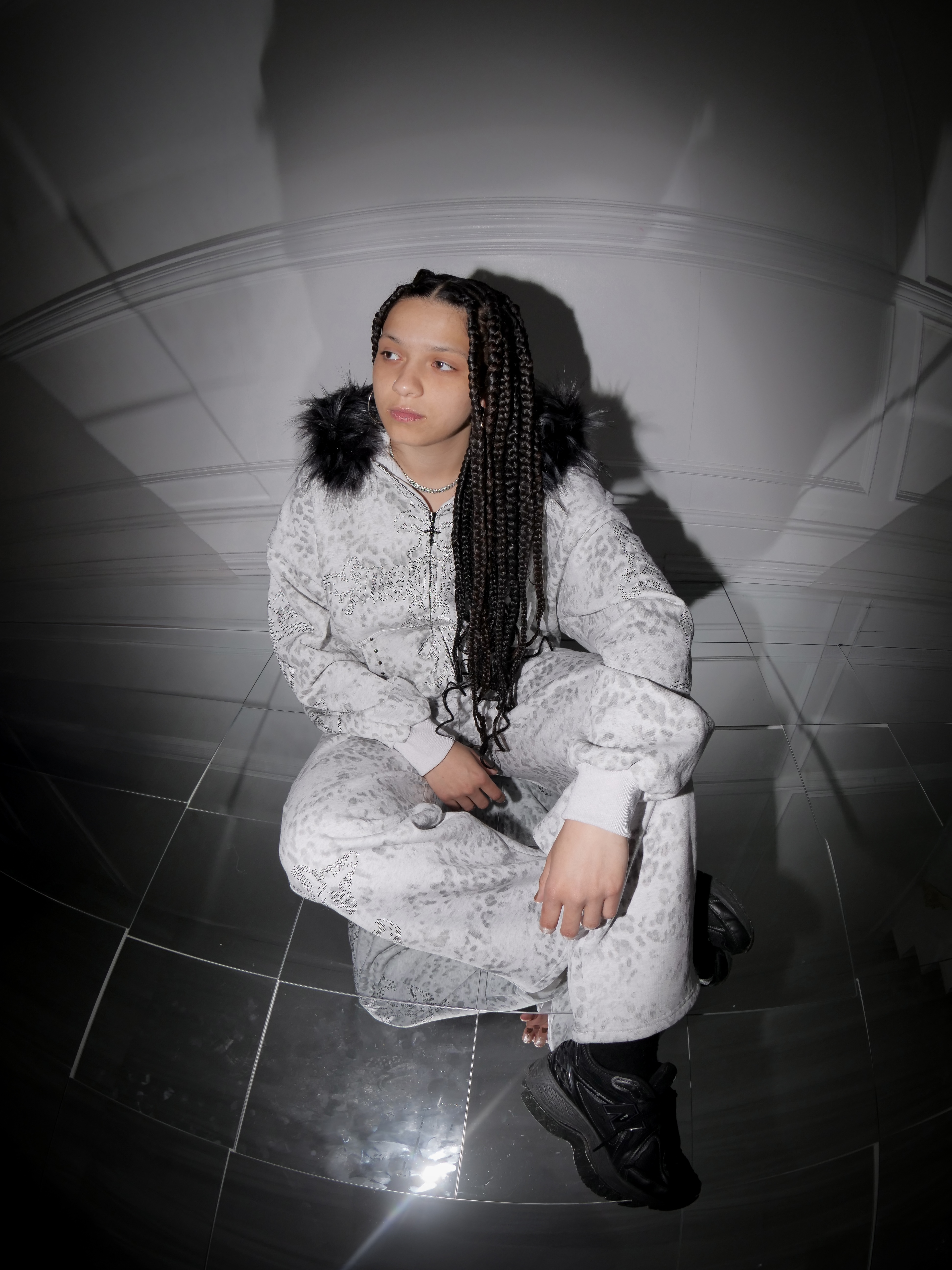 Person with long braided hair wearing white tracksuit with fur-trimmed hood, crouching on tiled floor in corner of room.