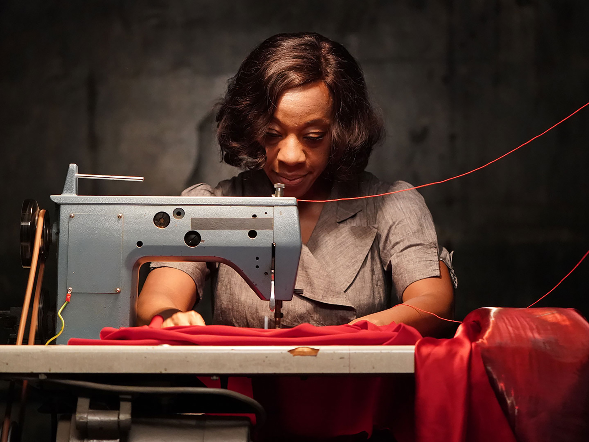 Woman operating a sewing machine, sewing red fabric on a dark background.