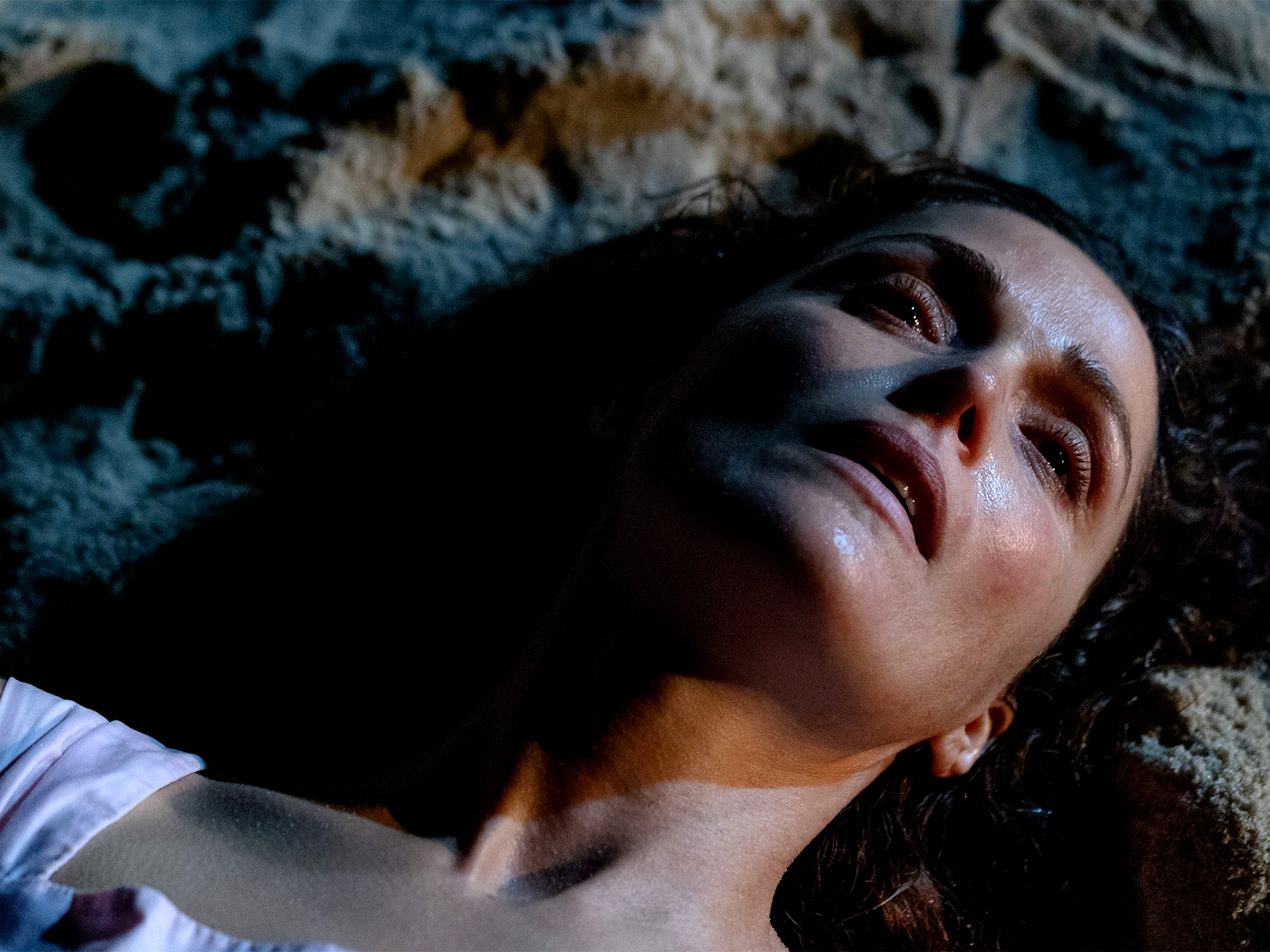 Woman with dark hair lying on rocky surface, eyes closed, wearing white top. Dramatic lighting creates shadows across face.
